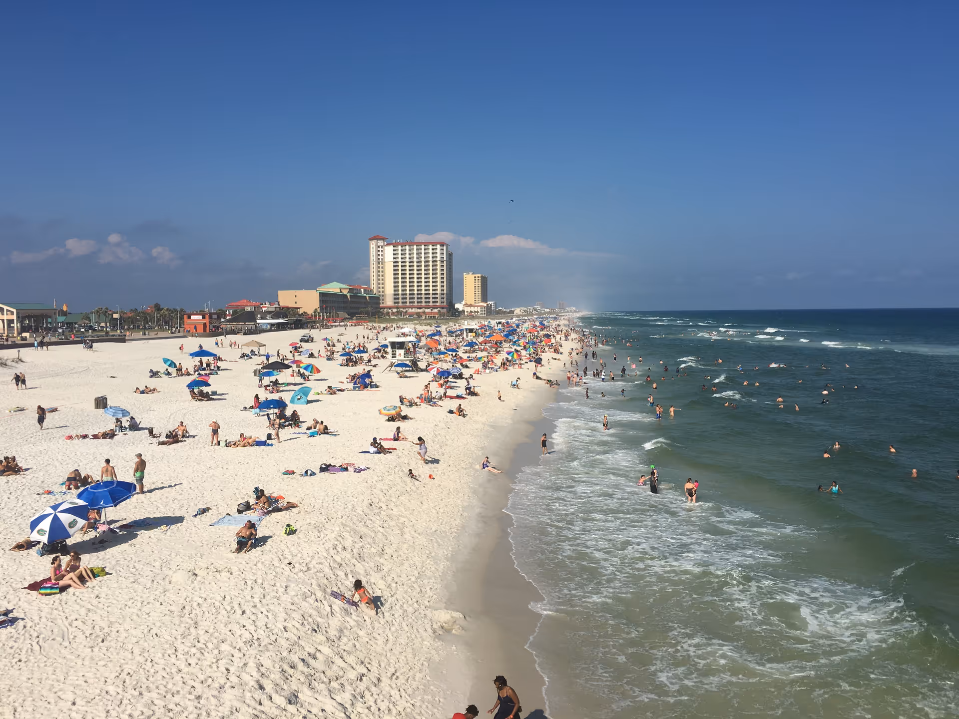 Busy sunny beach with people sunbathing along the shoreline and waves rolling in.