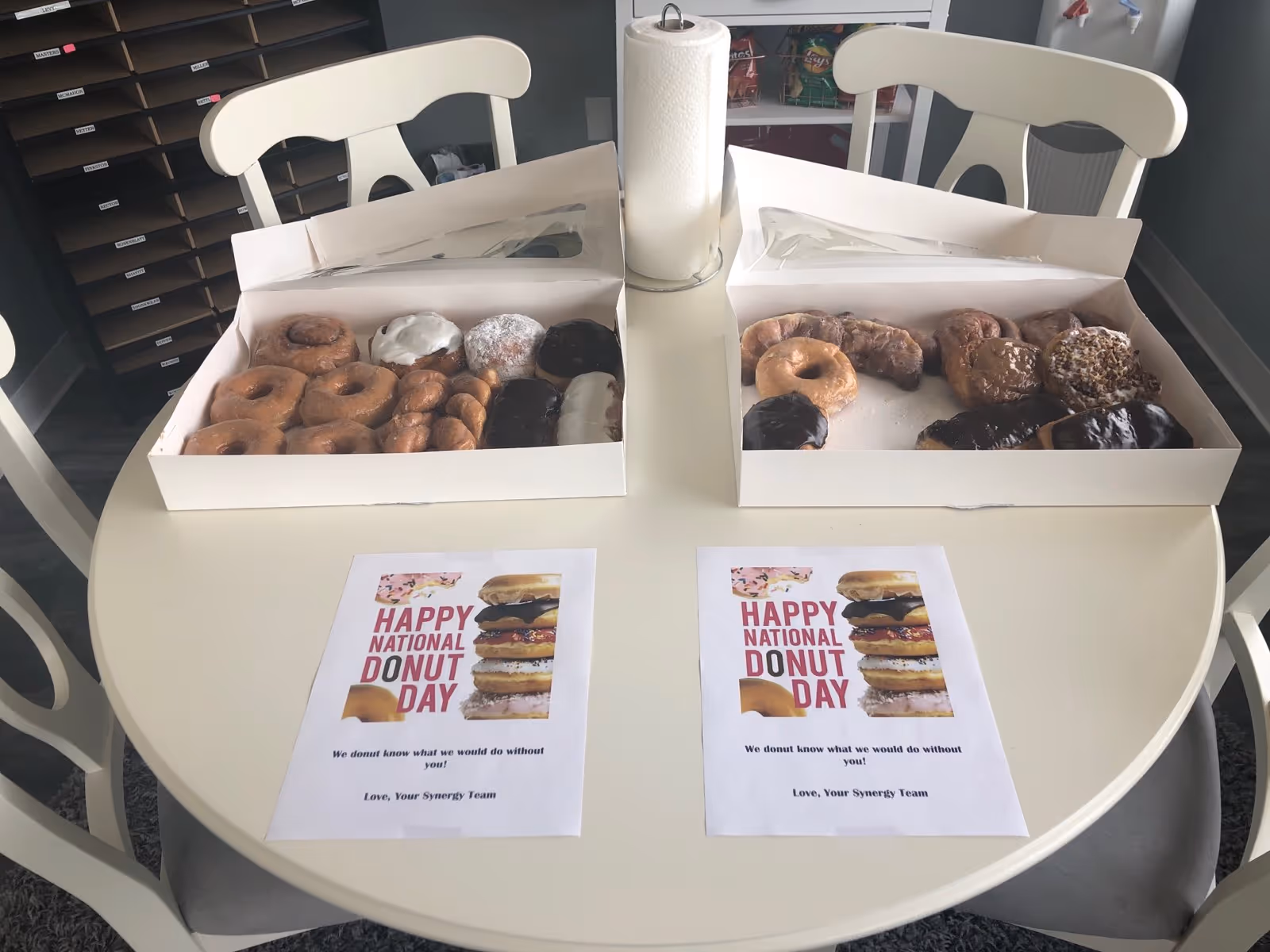 Two boxes of assorted donuts sit on a round white table beside National Donut Day flyers.