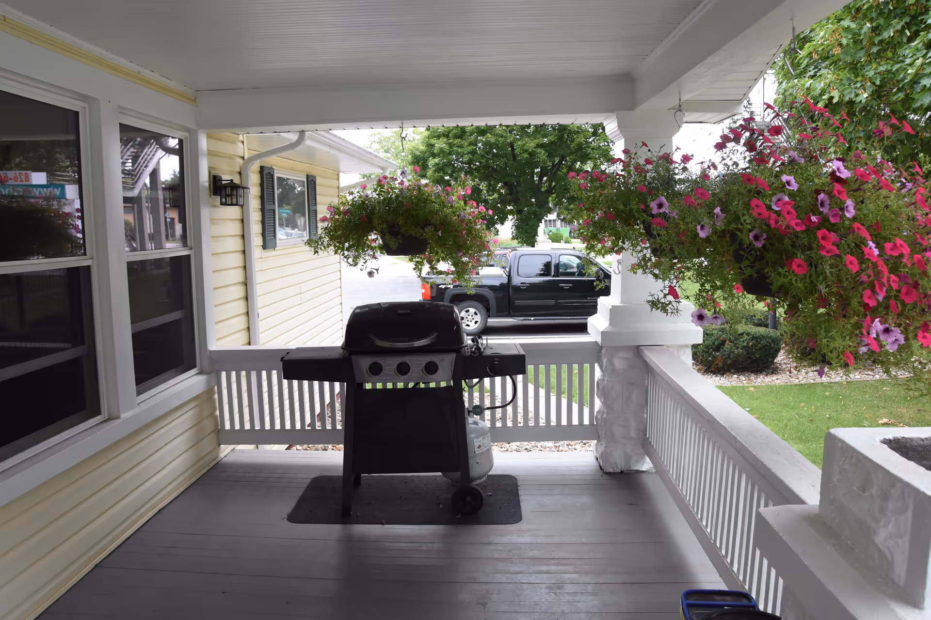 Covered porch with hanging flower baskets and a barbecue grill outside a yellow house.