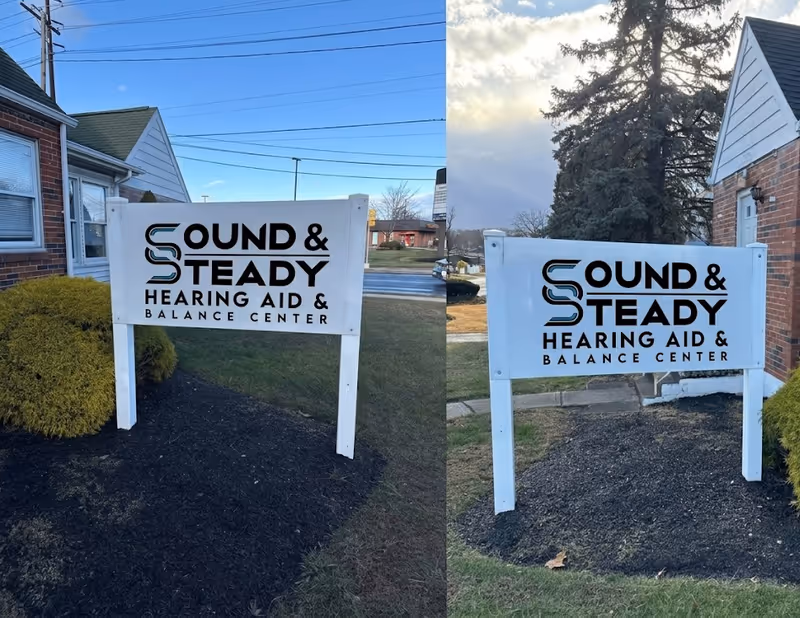 Two white signs display Sound and Steady Hearing Aid and Balance Center near a brick building.