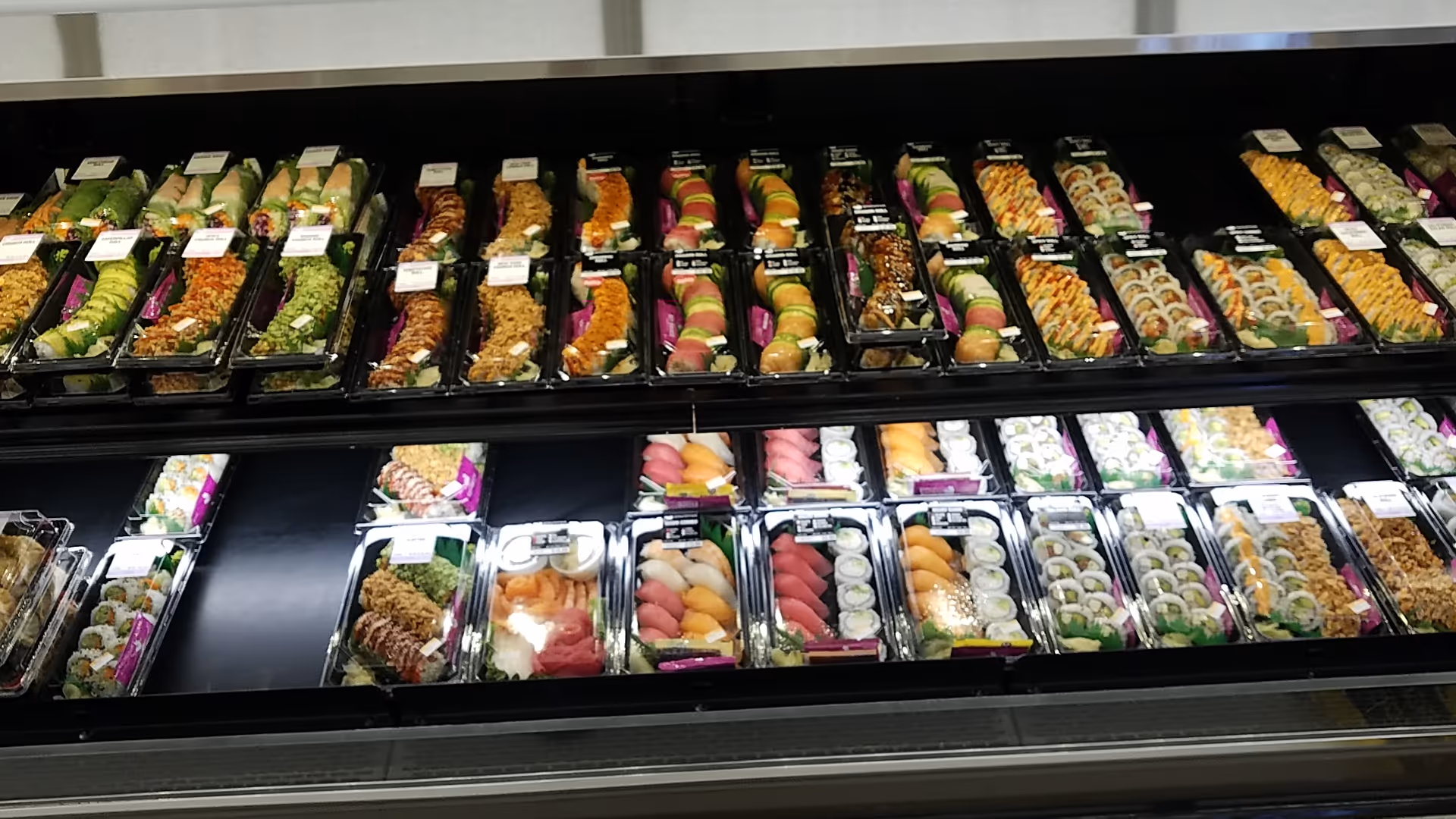 Rows of colorful sushi trays arranged in a refrigerated display case at a grocery store.