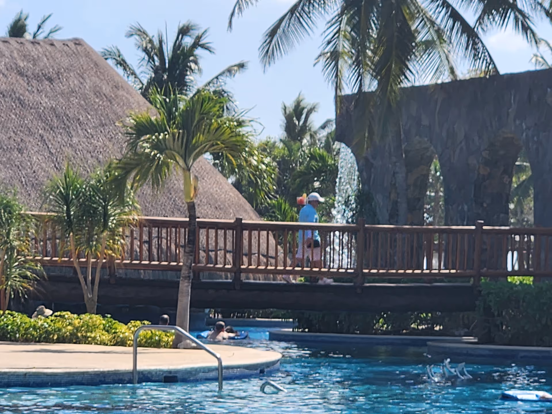 Resort-style outdoor pool area with a wooden bridge, palm trees, and people swimming.