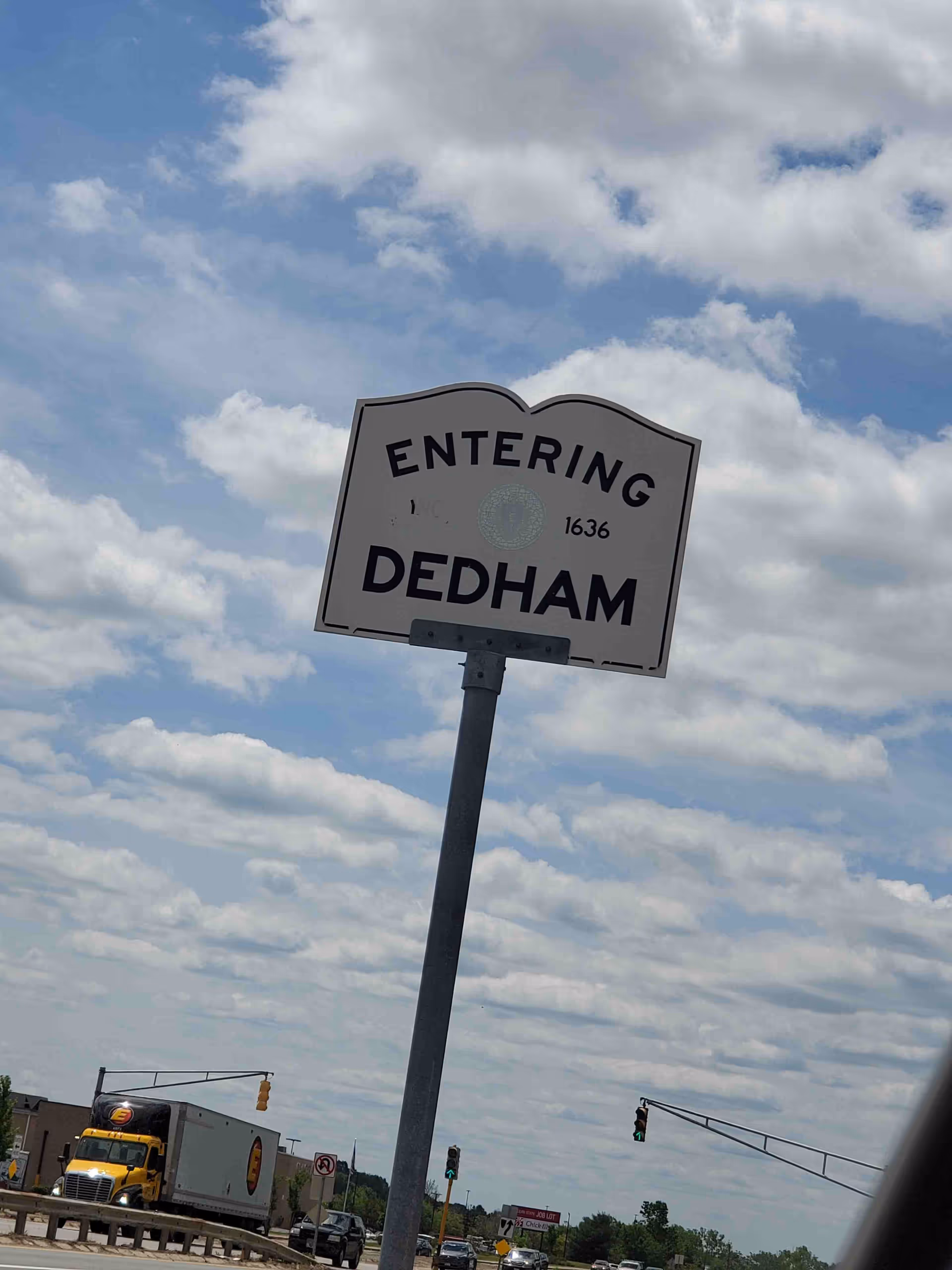 Large 'Entering Dedham' road sign against a blue sky with passing traffic.