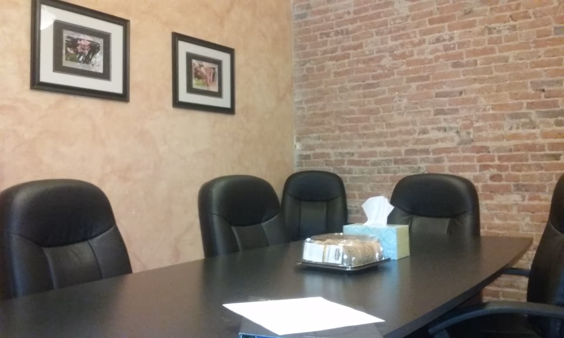 A conference-style interior with a long dark table, black leather chairs, brick wall backdrop, framed photos.