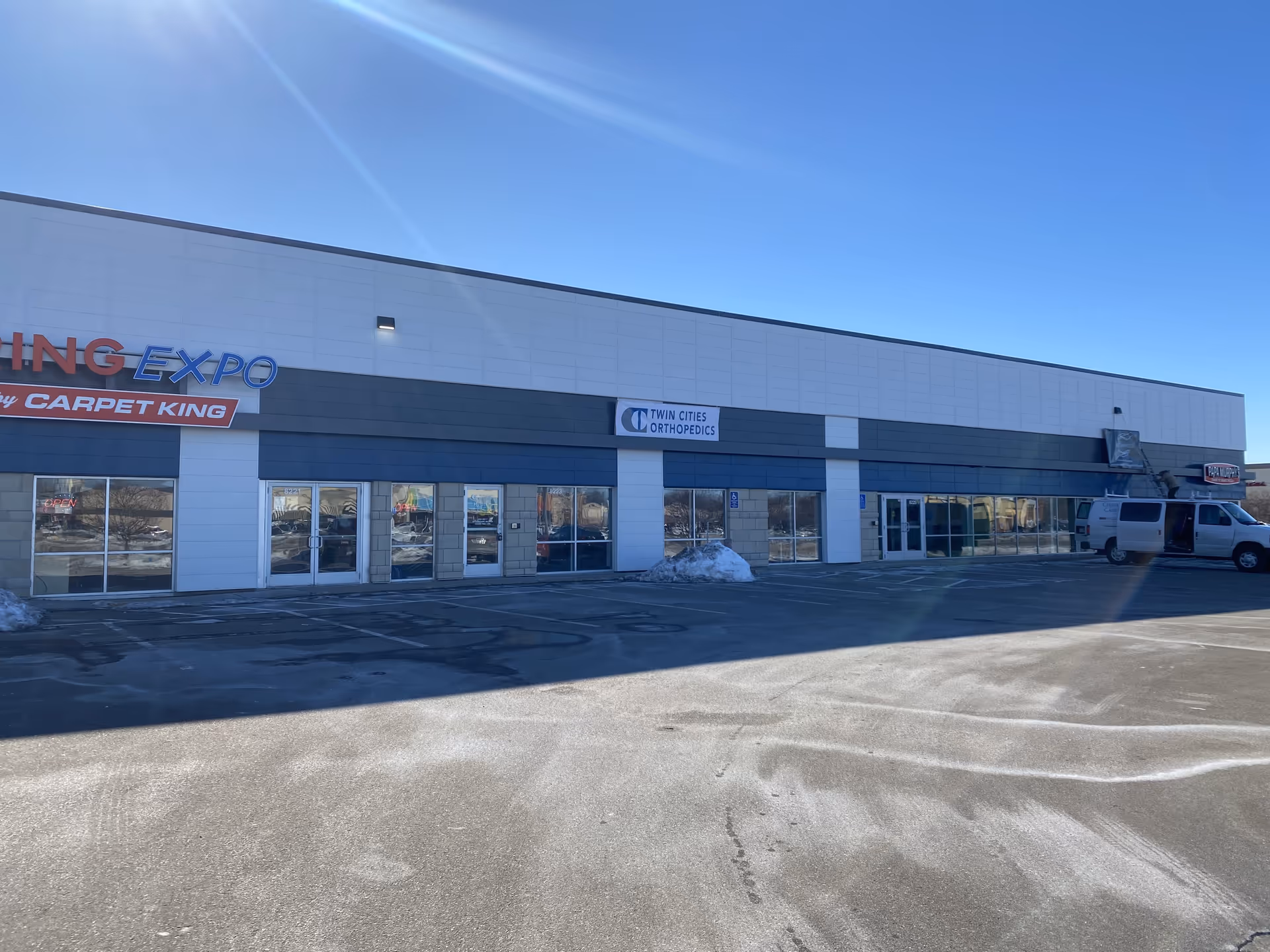 Long storefront building with multiple signs, glass doors, a white van, and a clear blue sky.