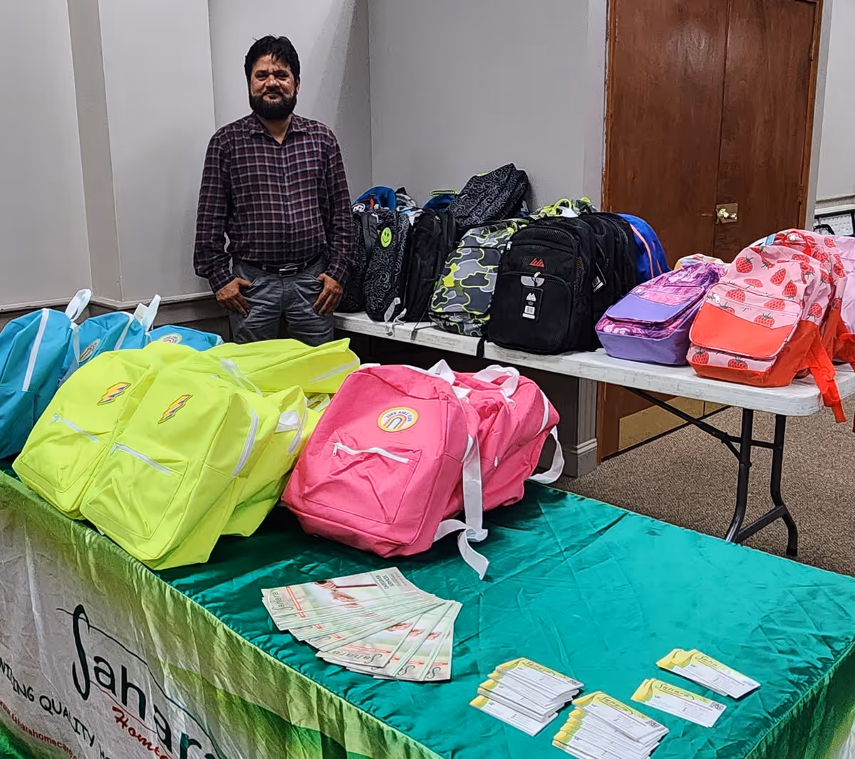A man stands beside a table covered with colorful backpacks indoors.