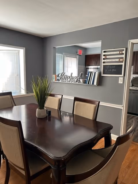 A dark wood dining table with beige chairs, plant centerpiece, and a kitchen pass-through sign.