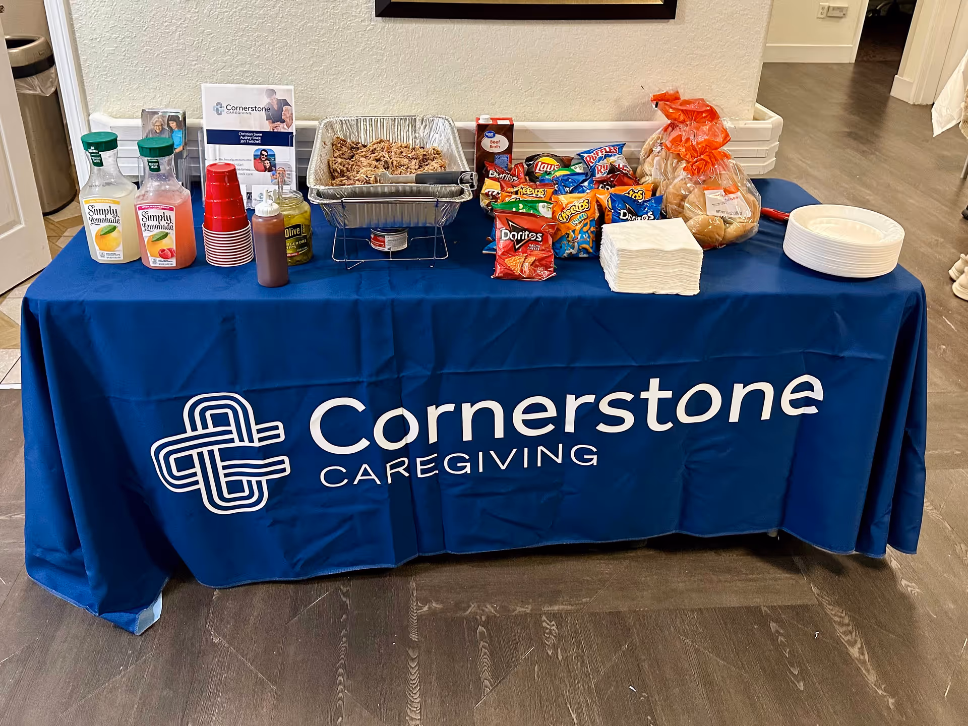 Indoor service table with snacks, drinks, napkins, and Cornerstone Caregiving branding on blue cloth