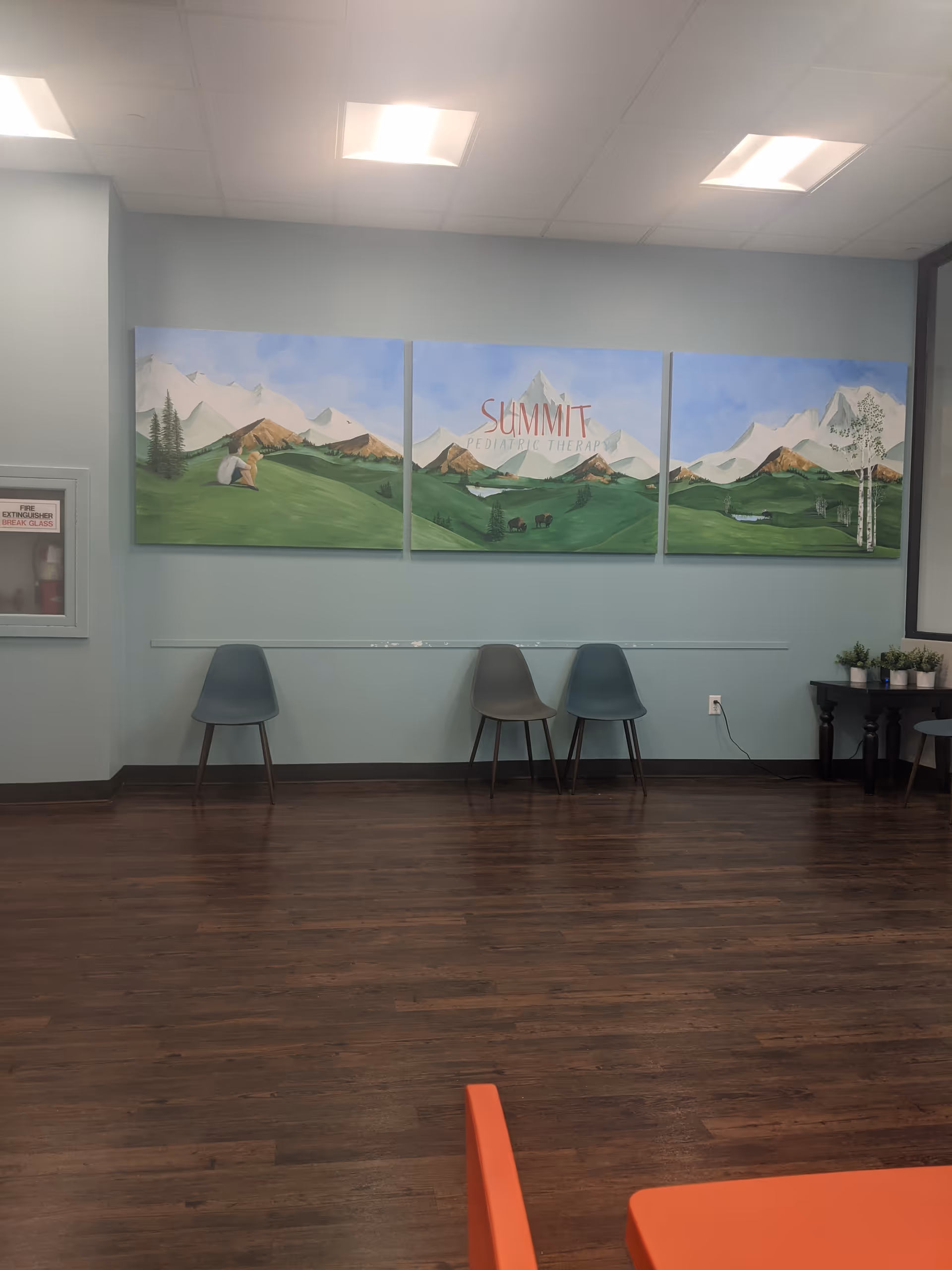 Interior waiting area with mountain mural, three chairs along a wall, wooden floor.