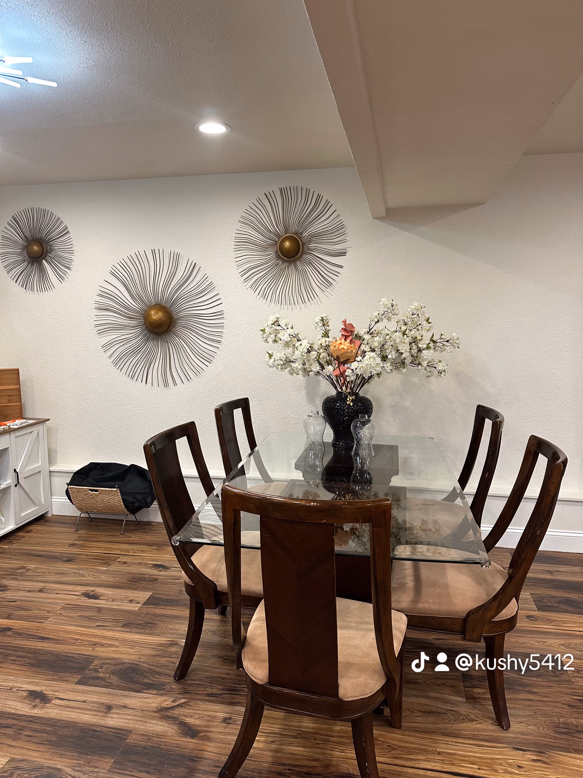 Glass dining table with six wooden chairs and floral centerpiece in a bright dining room.