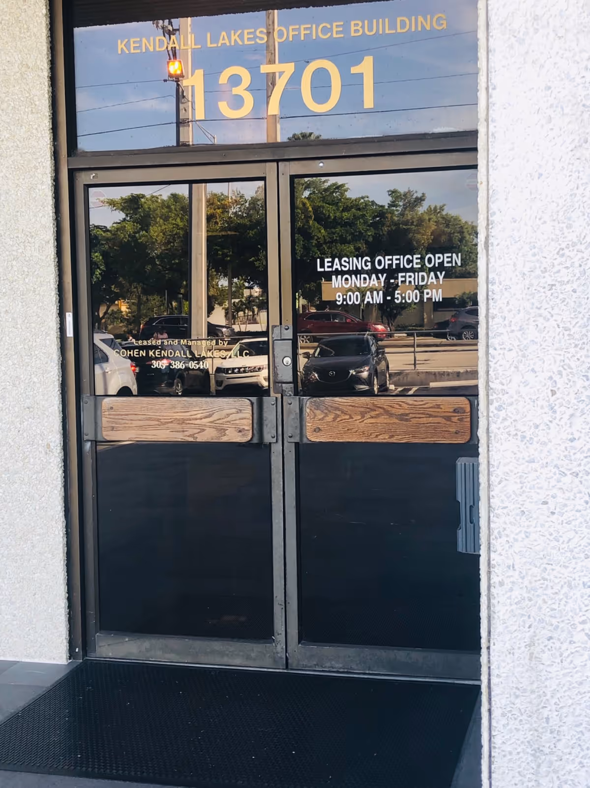 Glass doors show leasing office hours with reflections of parked cars outside.