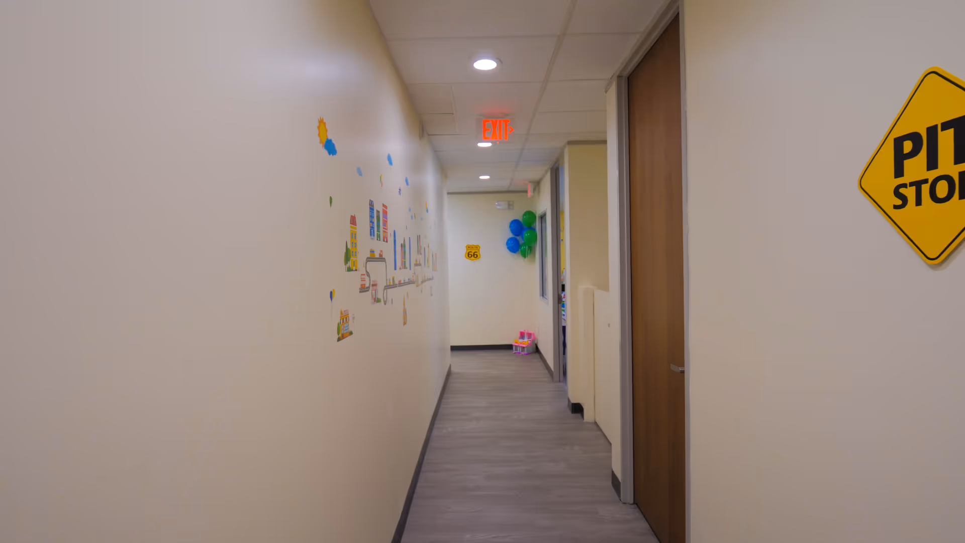 Long hallway in a facility with wall decorations, colorful decals, doors, and an EXIT sign.