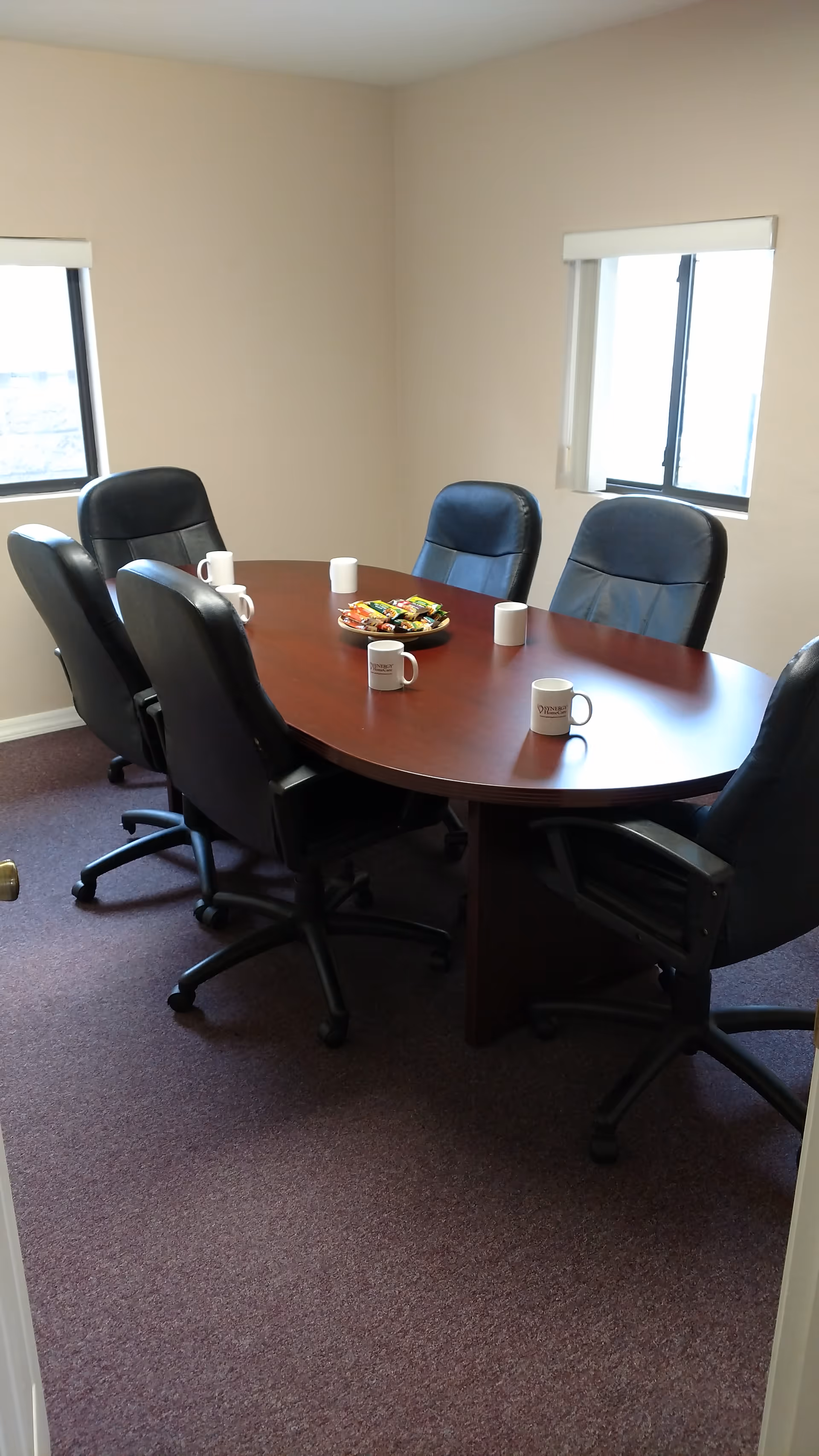 Round wooden conference table with six black chairs and mugs in a beige meeting room.