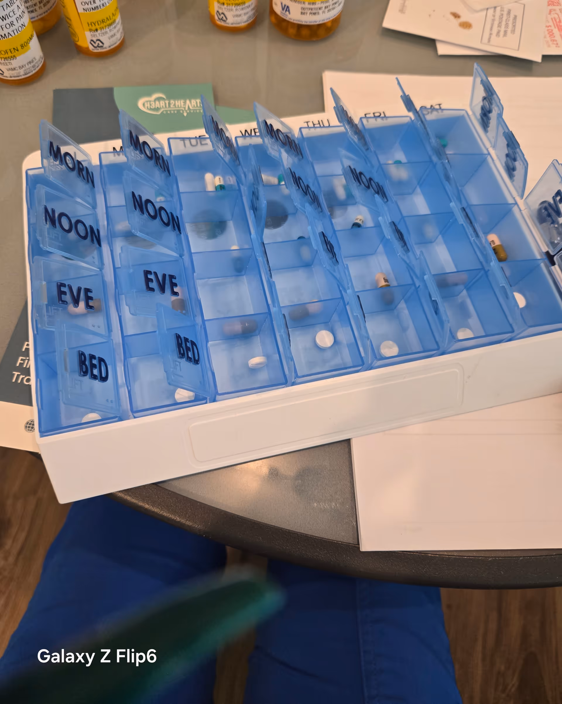 Blue weekly pill organizer with labeled compartments on a table beside medication bottles.