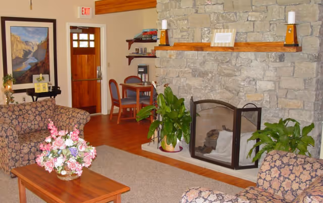 Cozy living room with a stone fireplace, floral sofa, plants, and coffee table; hospice setting.