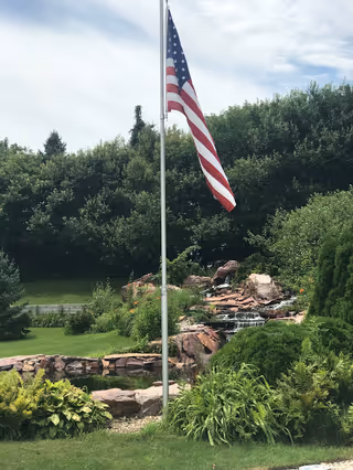 American flag on a tall pole beside a landscaped rock waterfall and greenery.