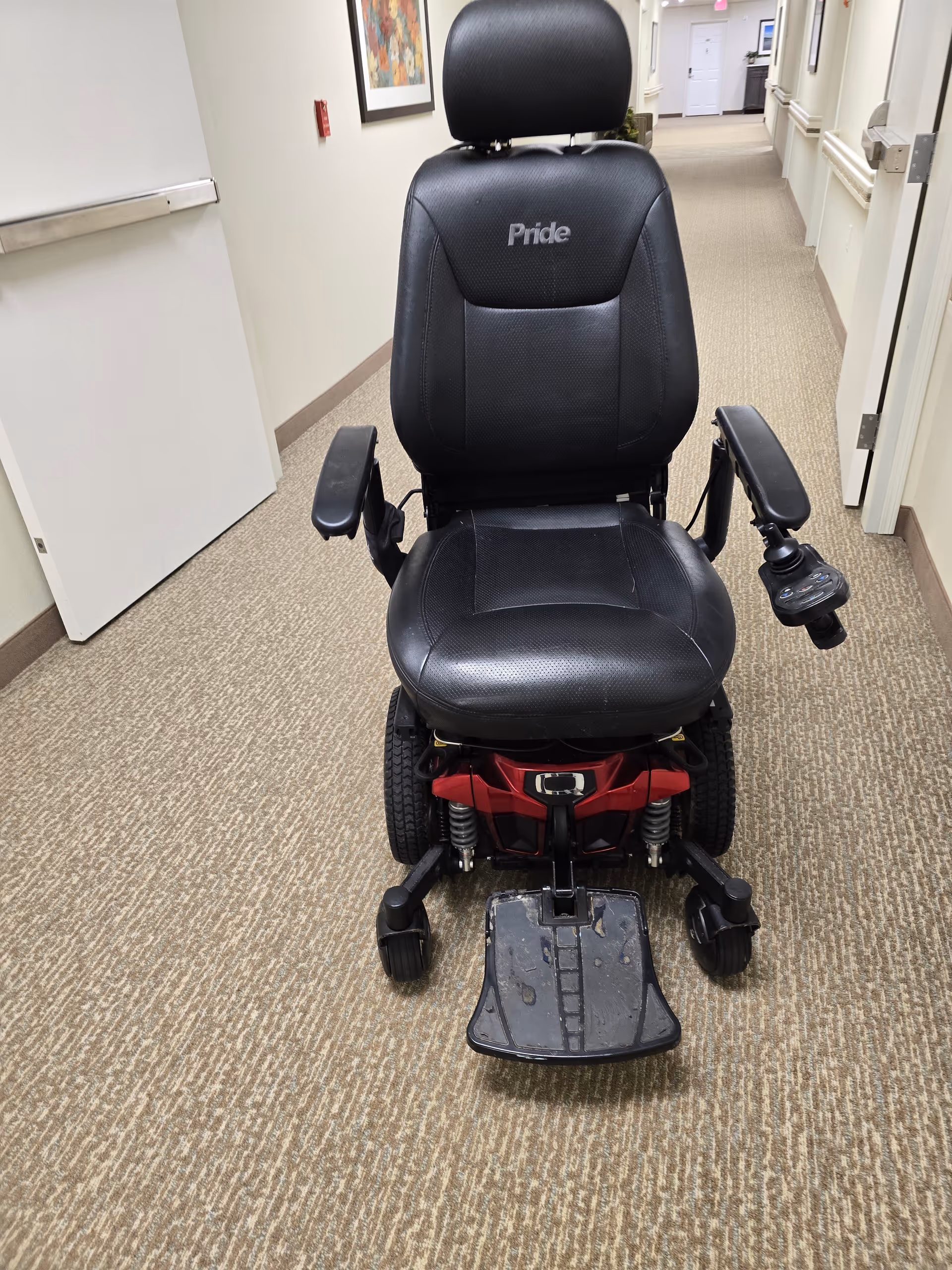 A black Pride mobility scooter sits in a carpeted hallway with doors and framed art.