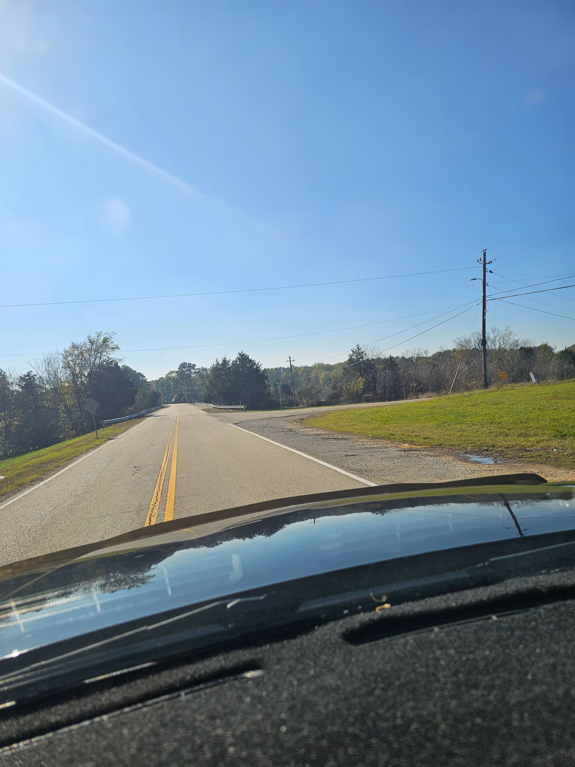 View from a car windshield of a straight rural road under a clear blue sky.