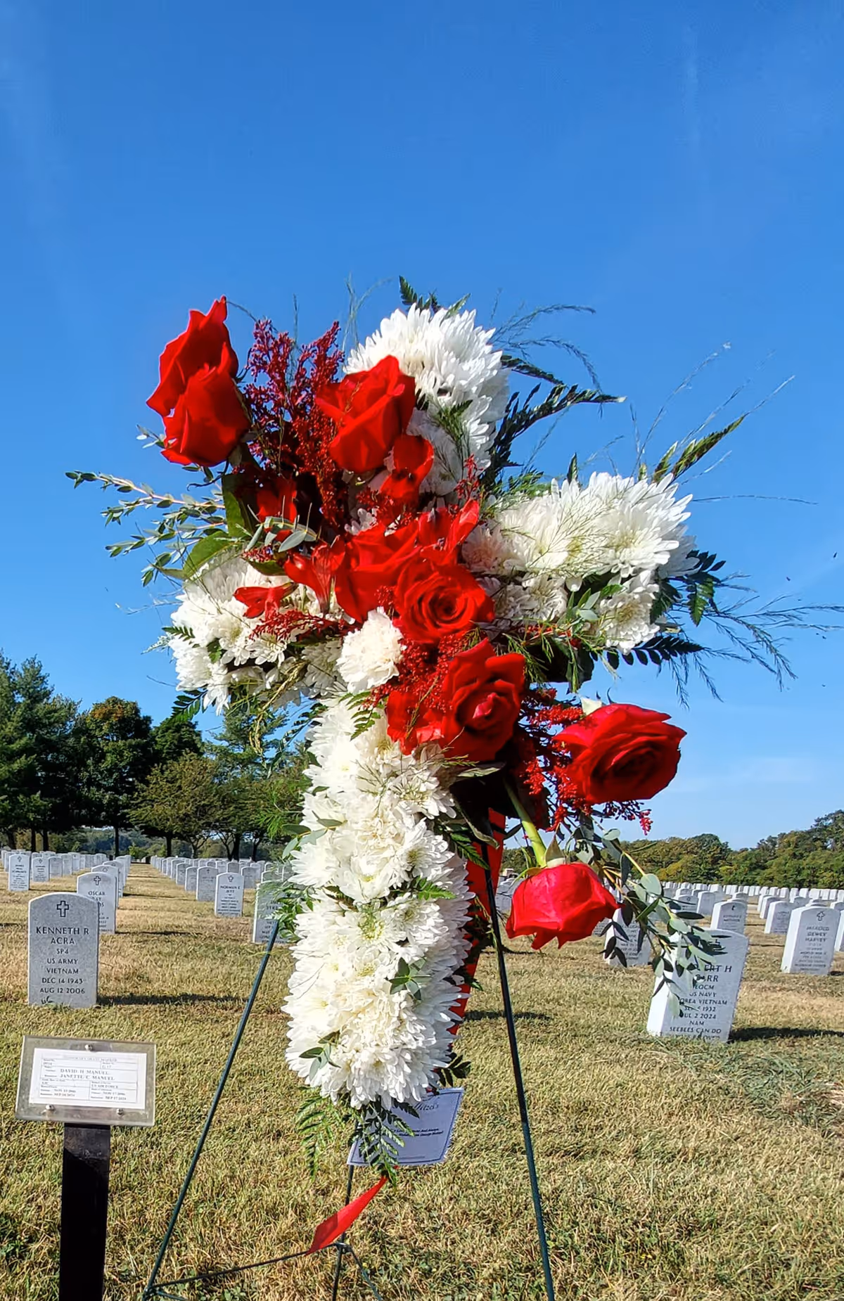 Large red and white floral arrangement on a tripod amid cemetery gravestones outdoors.