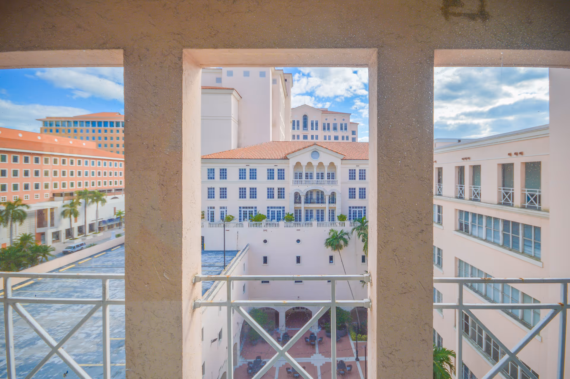 Balcony view overlooking a sunlit courtyard with white buildings and palm trees.