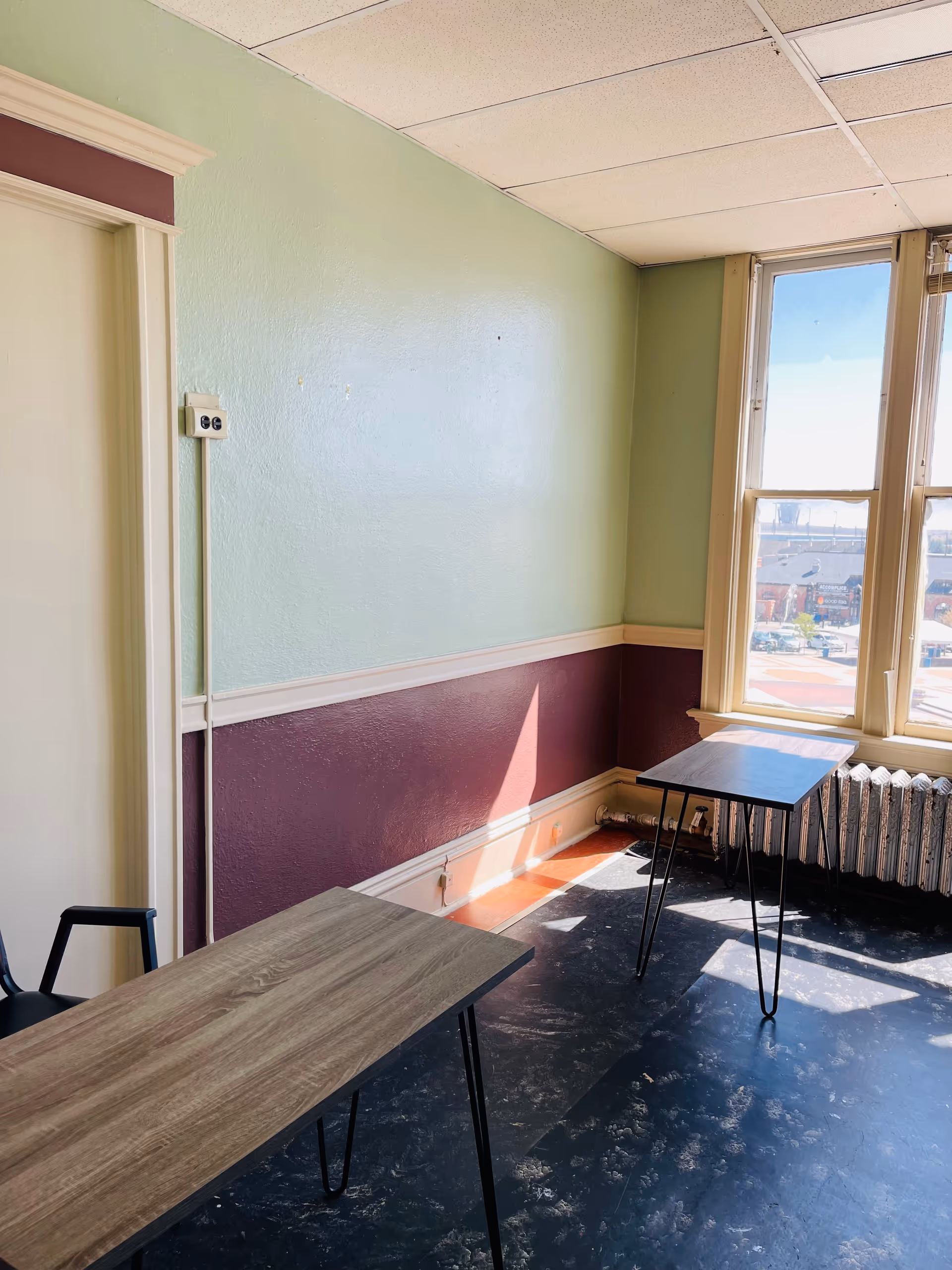 Sunlit corner room with pastel green walls, two wooden tables, and a radiator.