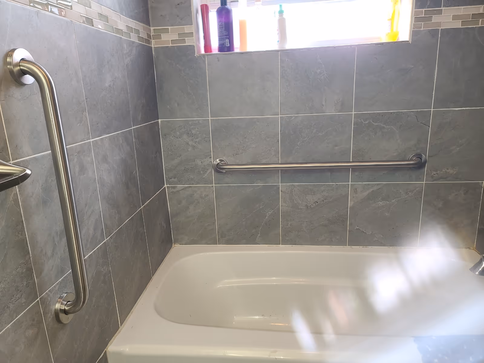 Grey tiled bathroom featuring a bathtub, wall grab bar, and safety features.