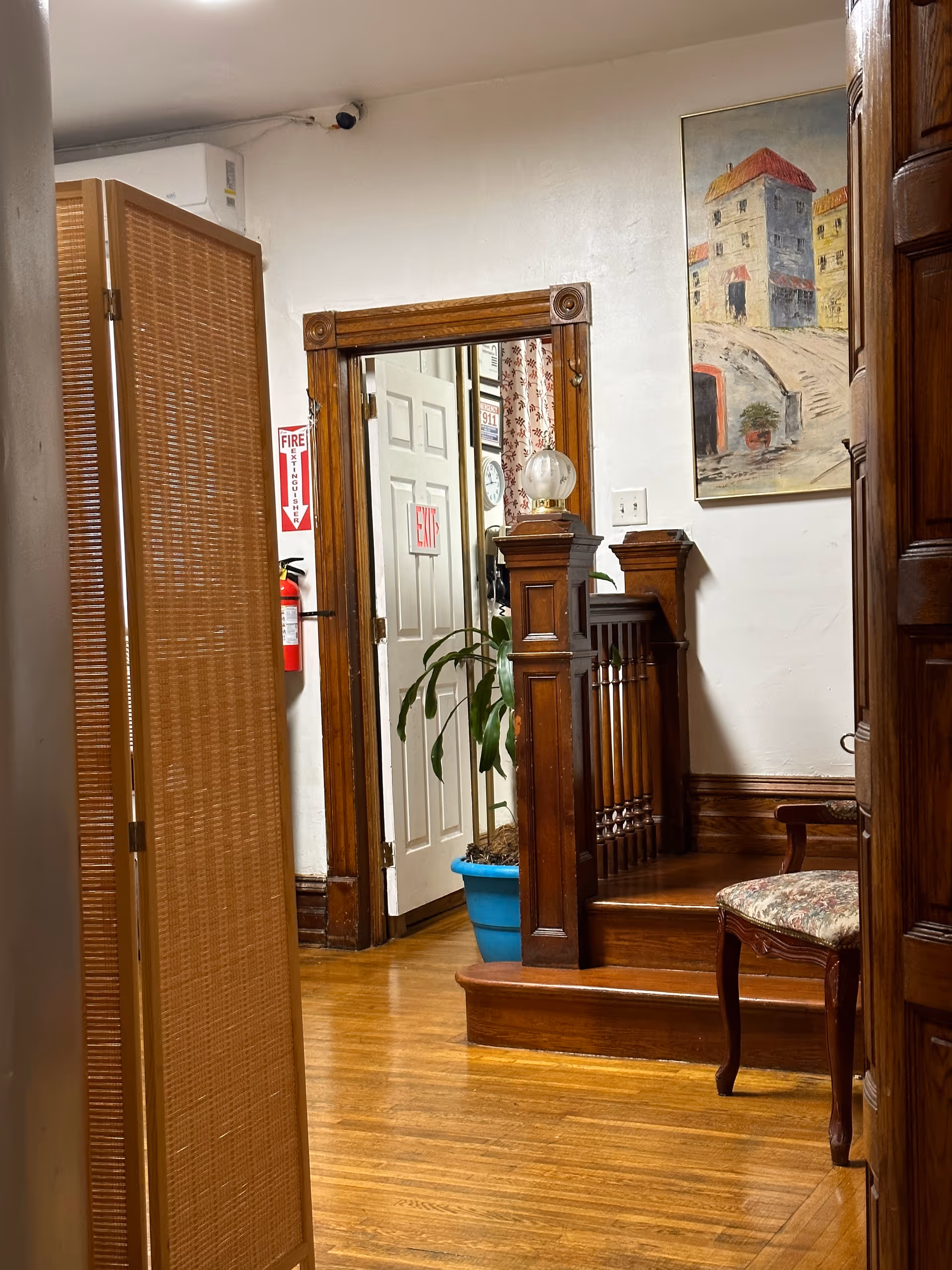 Interior hallway with wooden staircase, plant in blue pot, painting, and exit door.