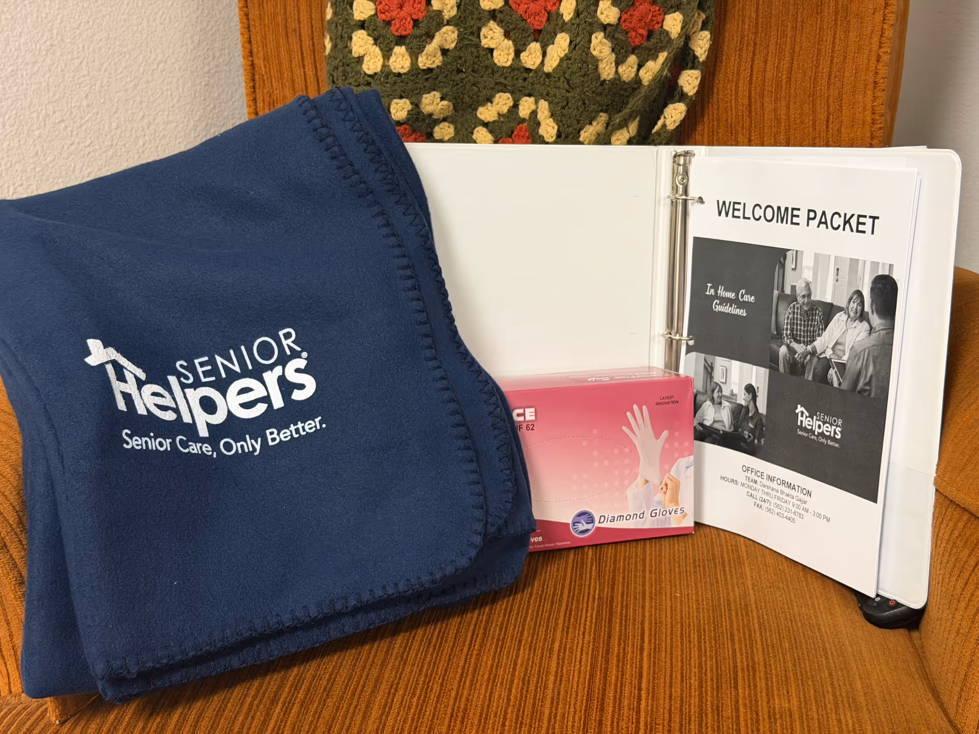 Blue Senior Helpers blanket, welcome packet binder, and box of gloves on a wooden bench.