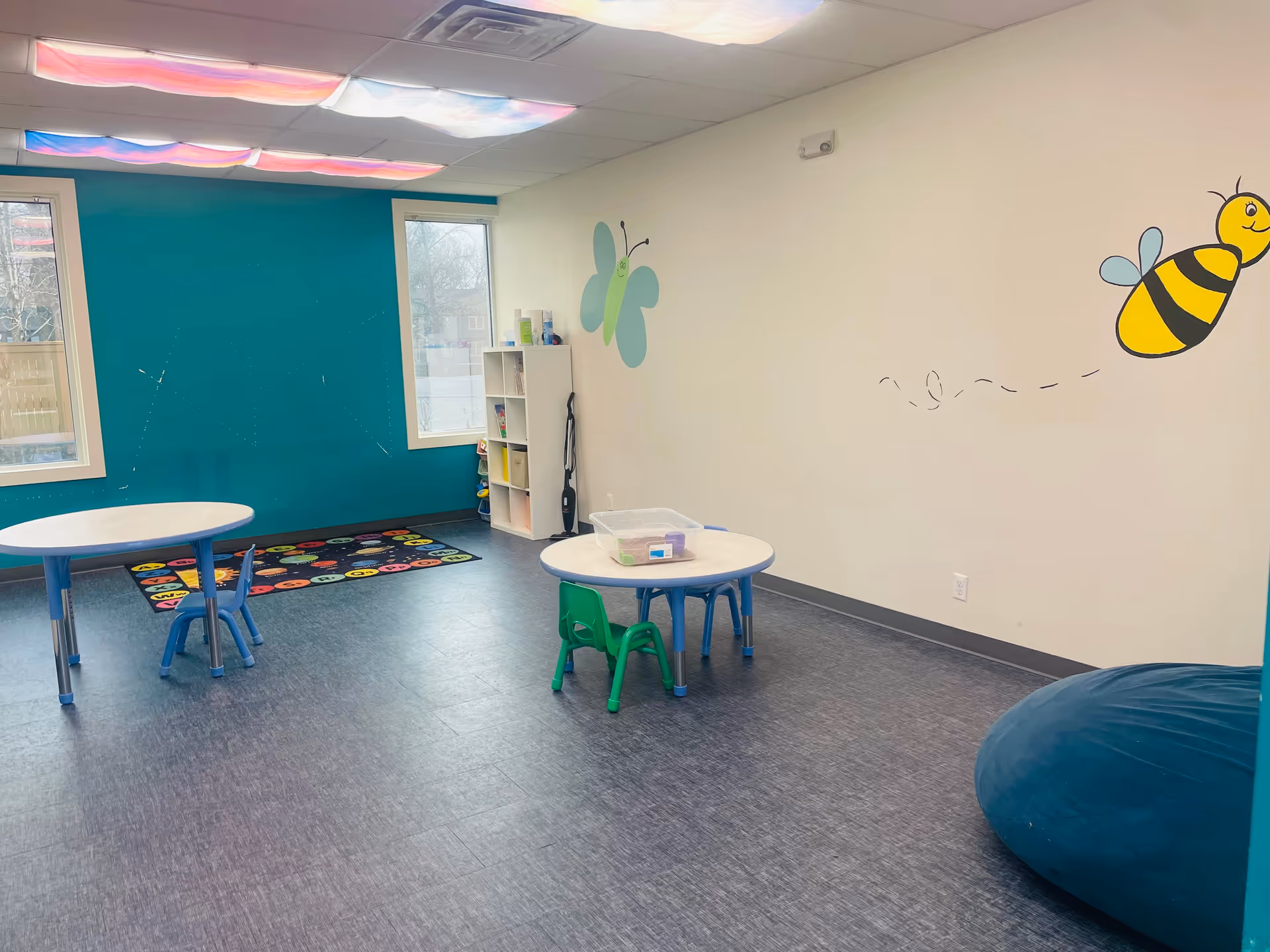 Colorful children's playroom with small tables, chairs, rug, and bee mural on walls.