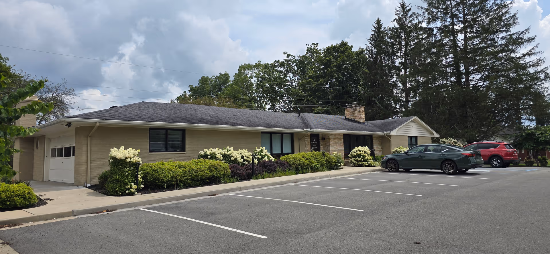 Single-story beige building with a parking lot and landscaped shrubs in front.