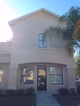 Two-story beige senior living building entrance with large windows and palm trees.