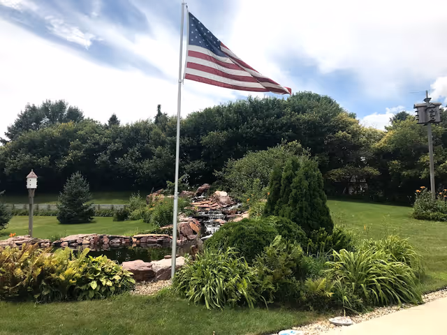 American flag on a tall pole overlooks a landscaped garden with rocks and a small waterfall.