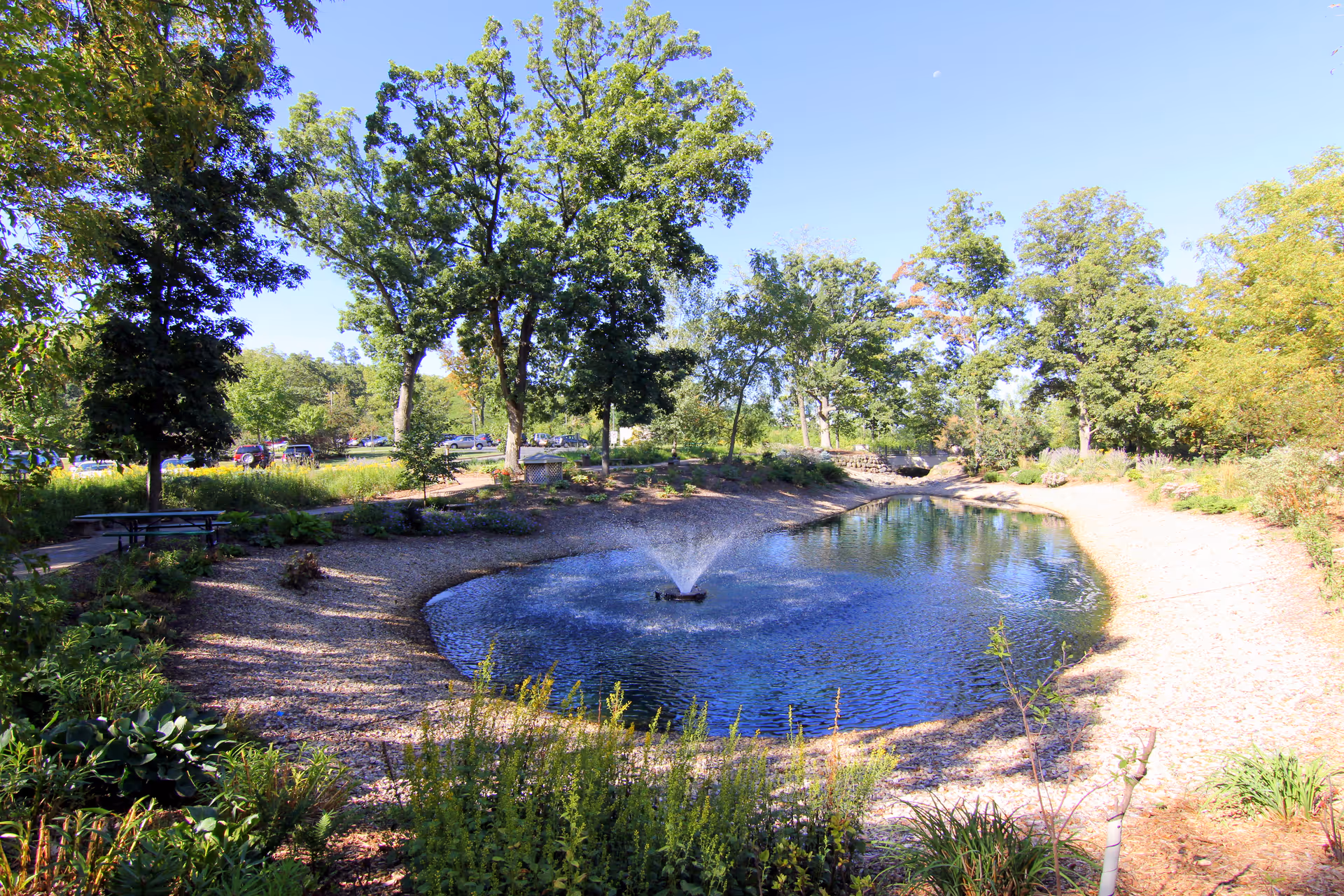 A landscaped outdoor park with a small pond, fountain, trees, and gravel paths.