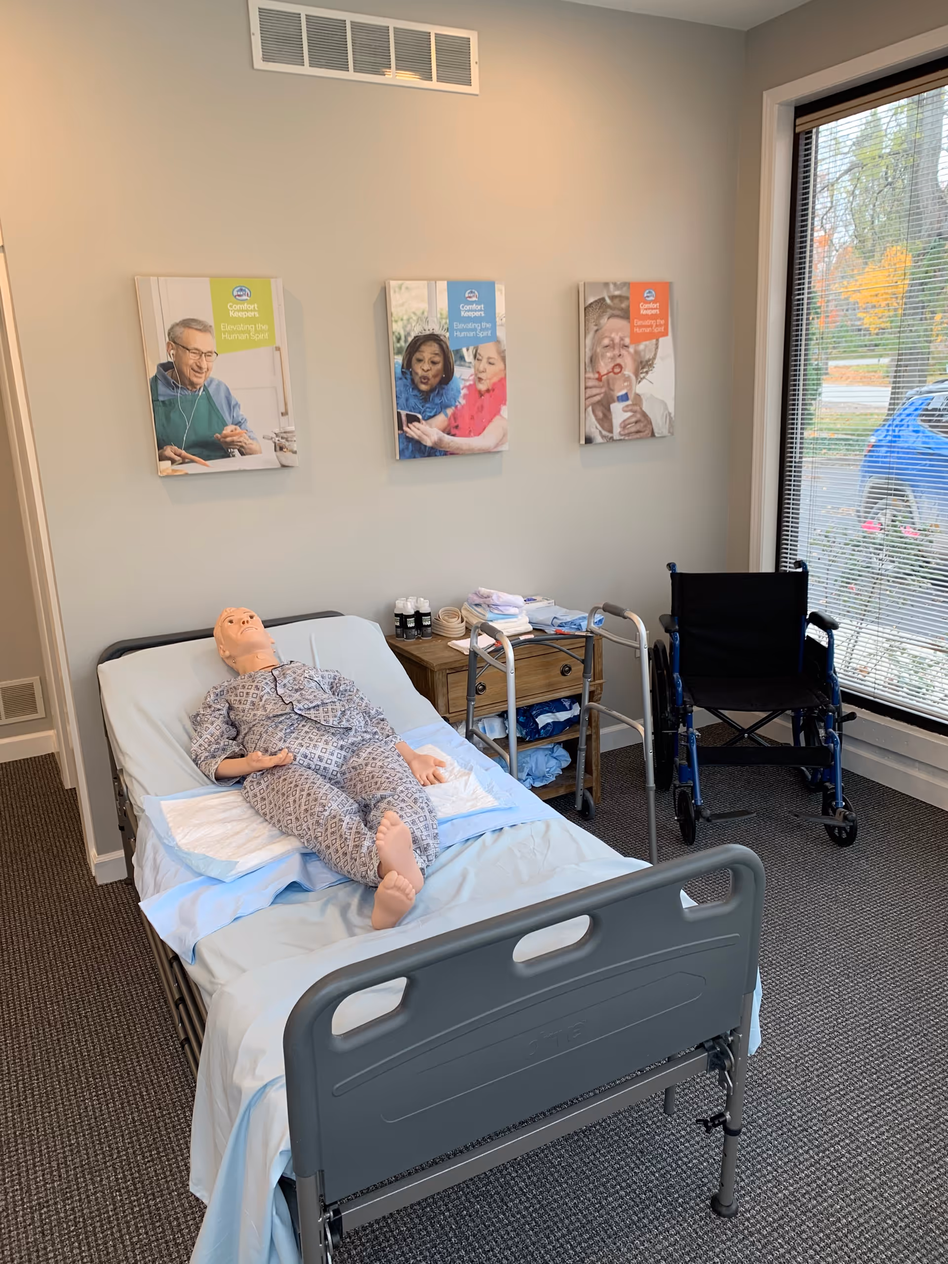 Hospital-style bed with a mannequin patient in a care room, wheelchair nearby, posters on the wall.