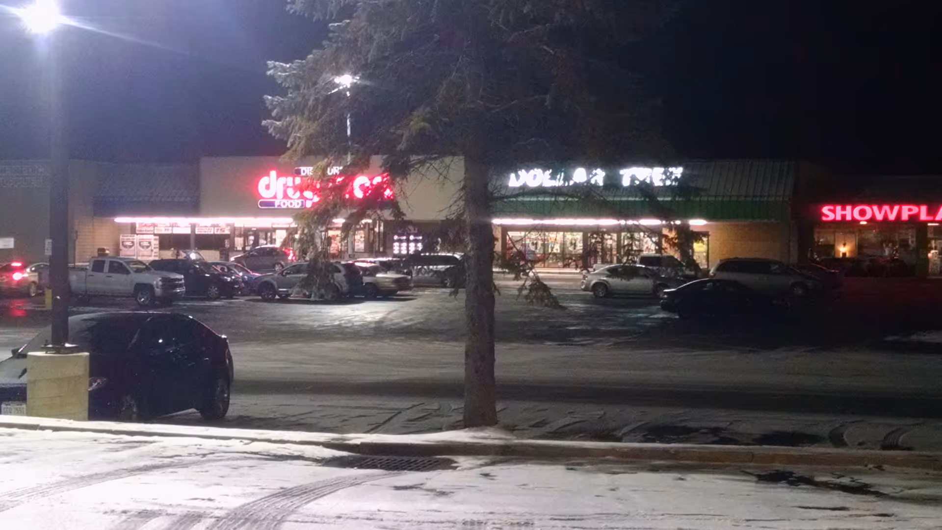 Nighttime exterior of a strip mall with Discount Drug Mart, parked cars, and lit storefronts.
