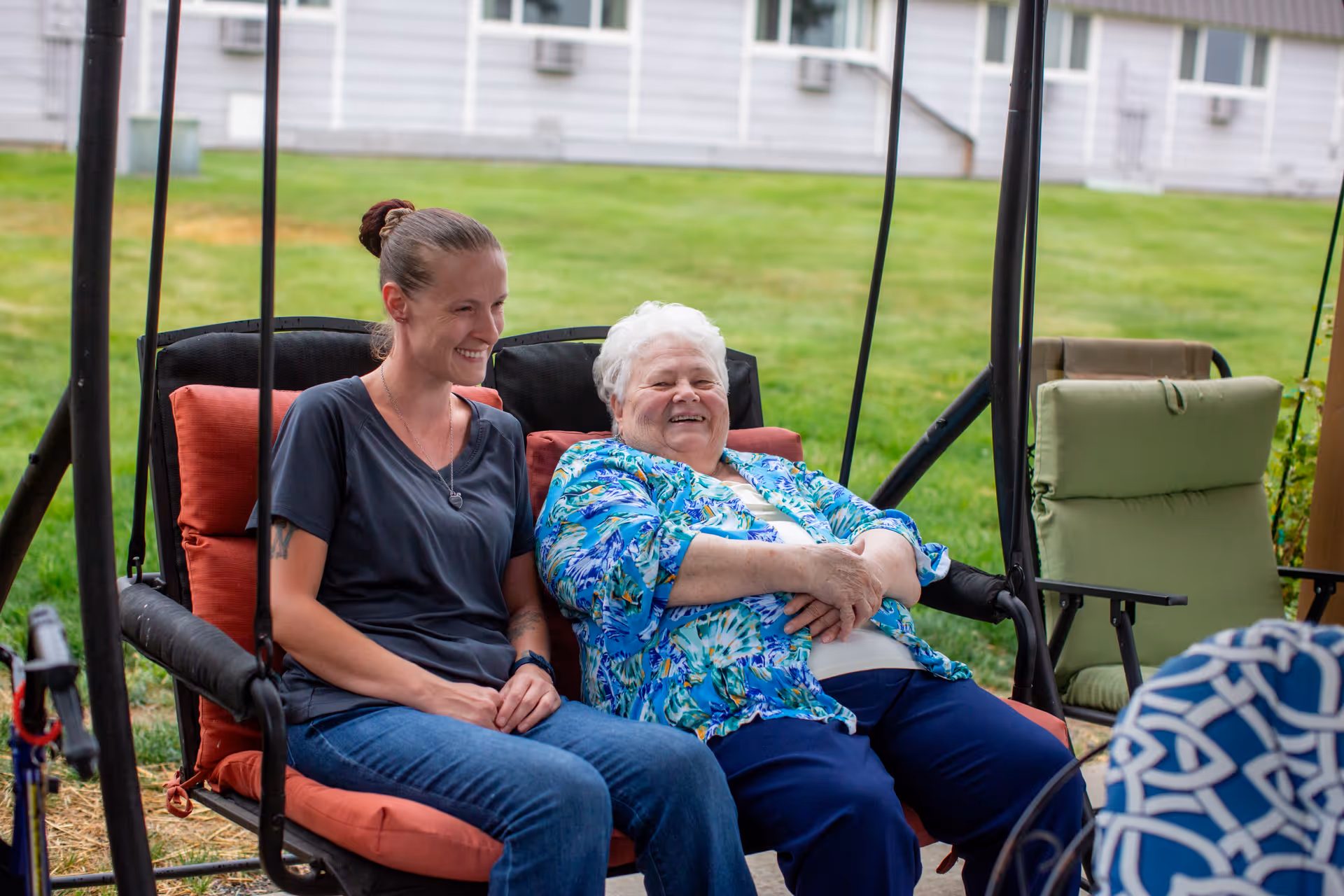 Two women sit on a red cushioned outdoor swing, smiling in a sunny facility yard.