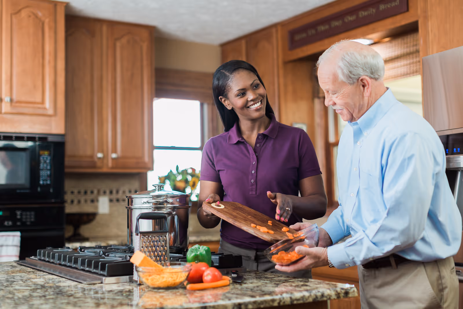 A caregiver and elderly man prepare vegetables together in a bright kitchen.