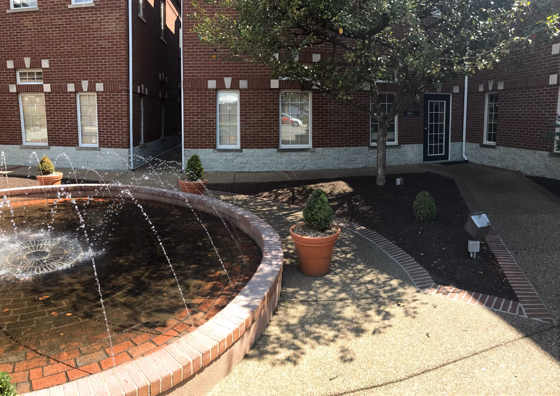 Brick building courtyard featuring a circular brick fountain, potted shrubs, and a tree near the entrance.