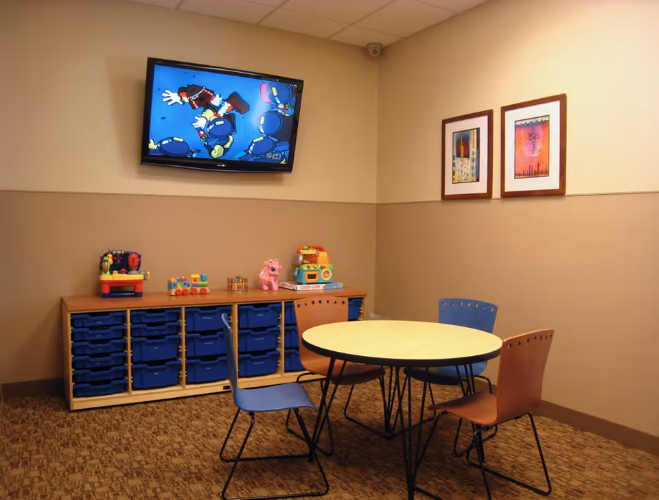 Play area with round table, colorful chairs, toy shelves, and a wall-mounted TV.