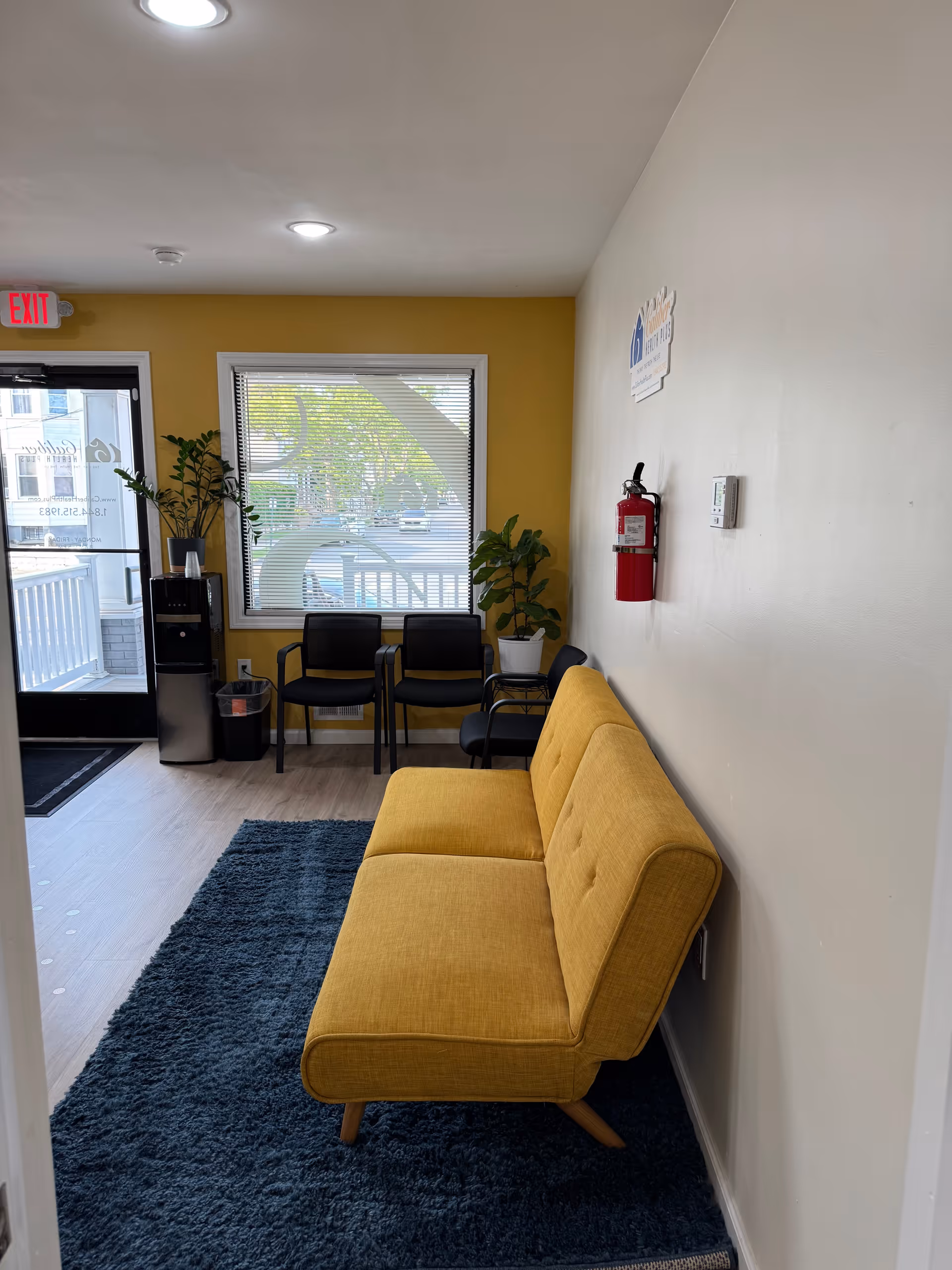 Bright interior waiting area with a yellow sofa, black chairs, plants, and an exit door.