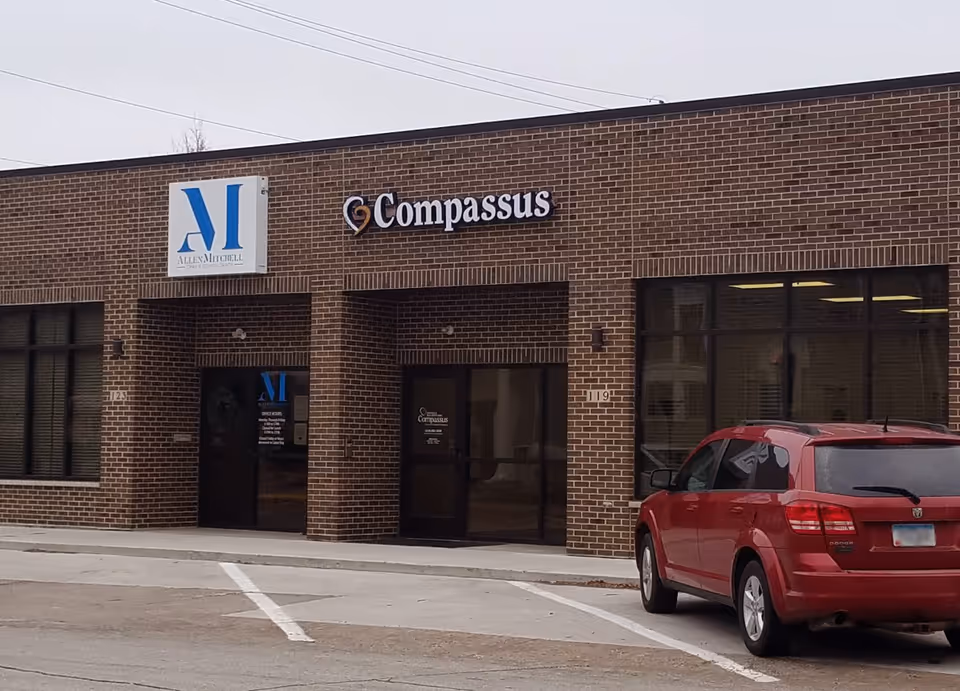 Brick building storefront displaying Compassus sign with a red SUV in a parking lot.