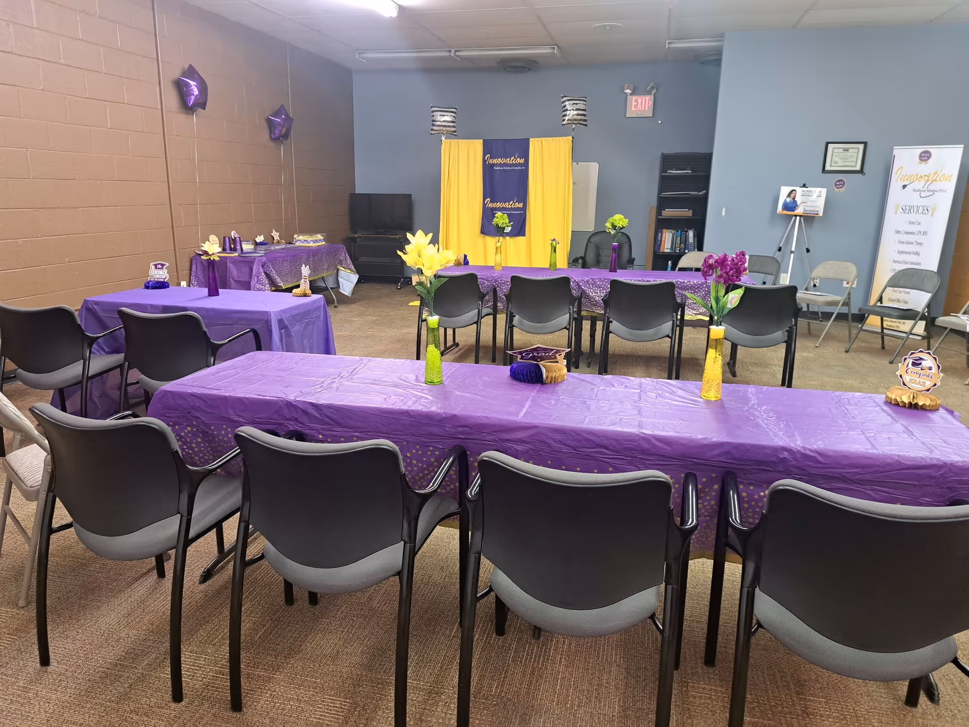 Indoor event room with purple tablecloths, chairs, and a stage backdrop.