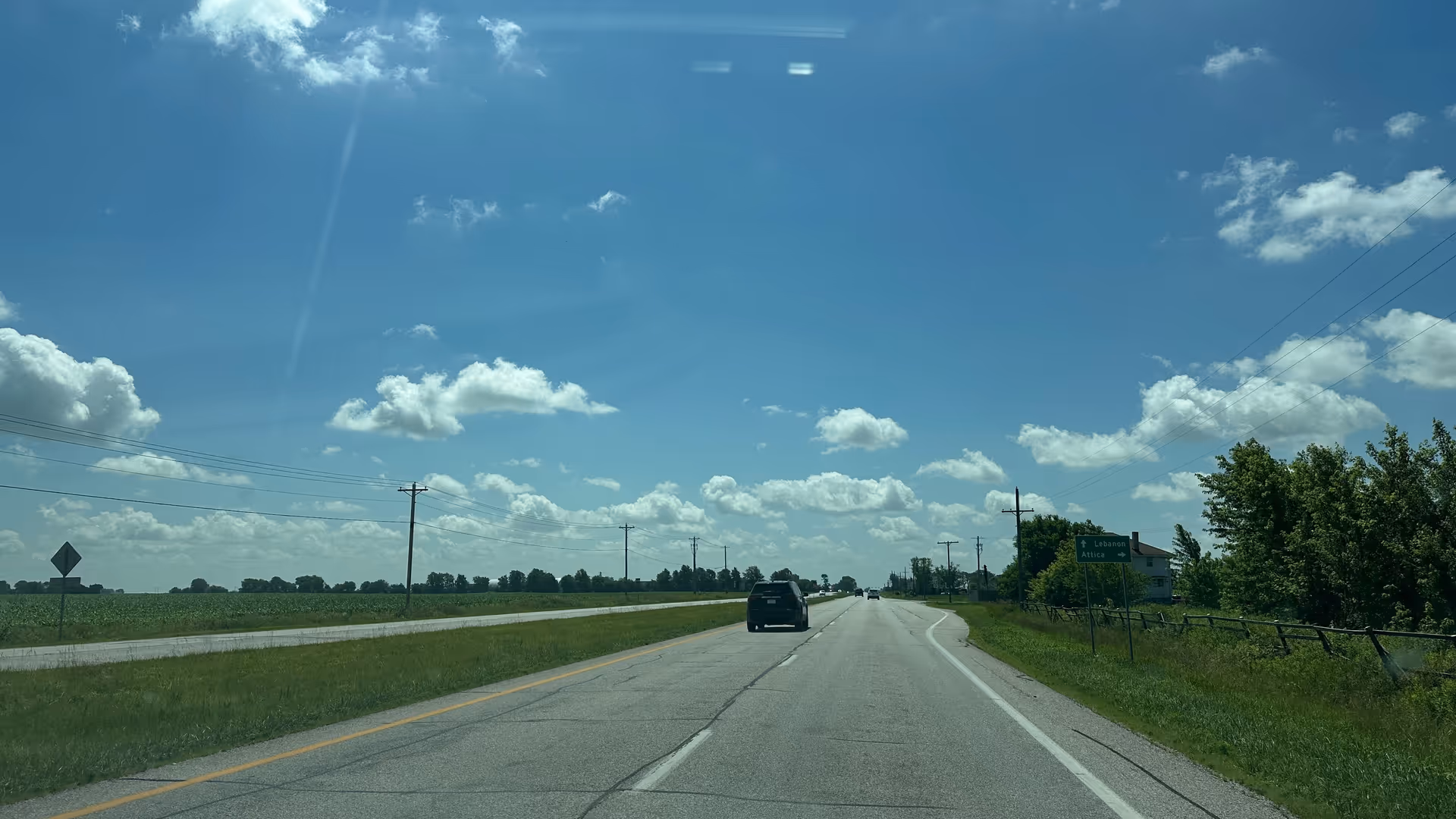 Long rural road flanked by fields and trees under a blue sky.