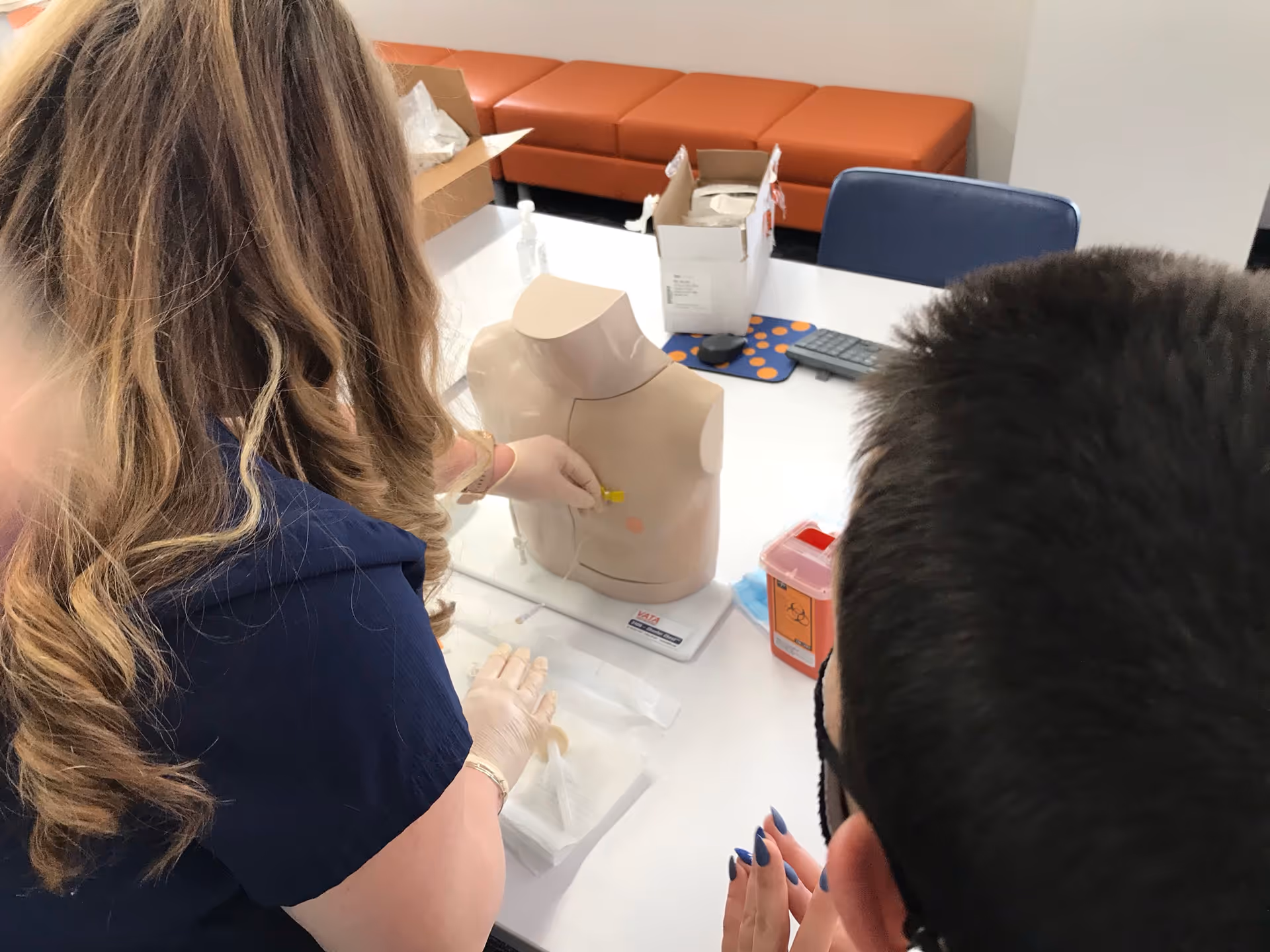 Healthcare workers practice on a medical mannequin at a table in a training room.