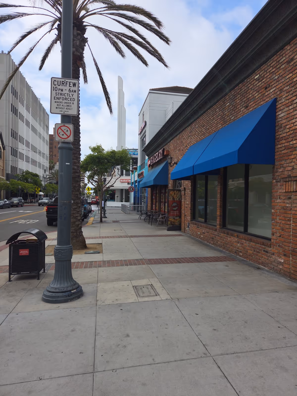Urban street view with brick storefronts, blue awnings, palm trees, and a wide sidewalk.