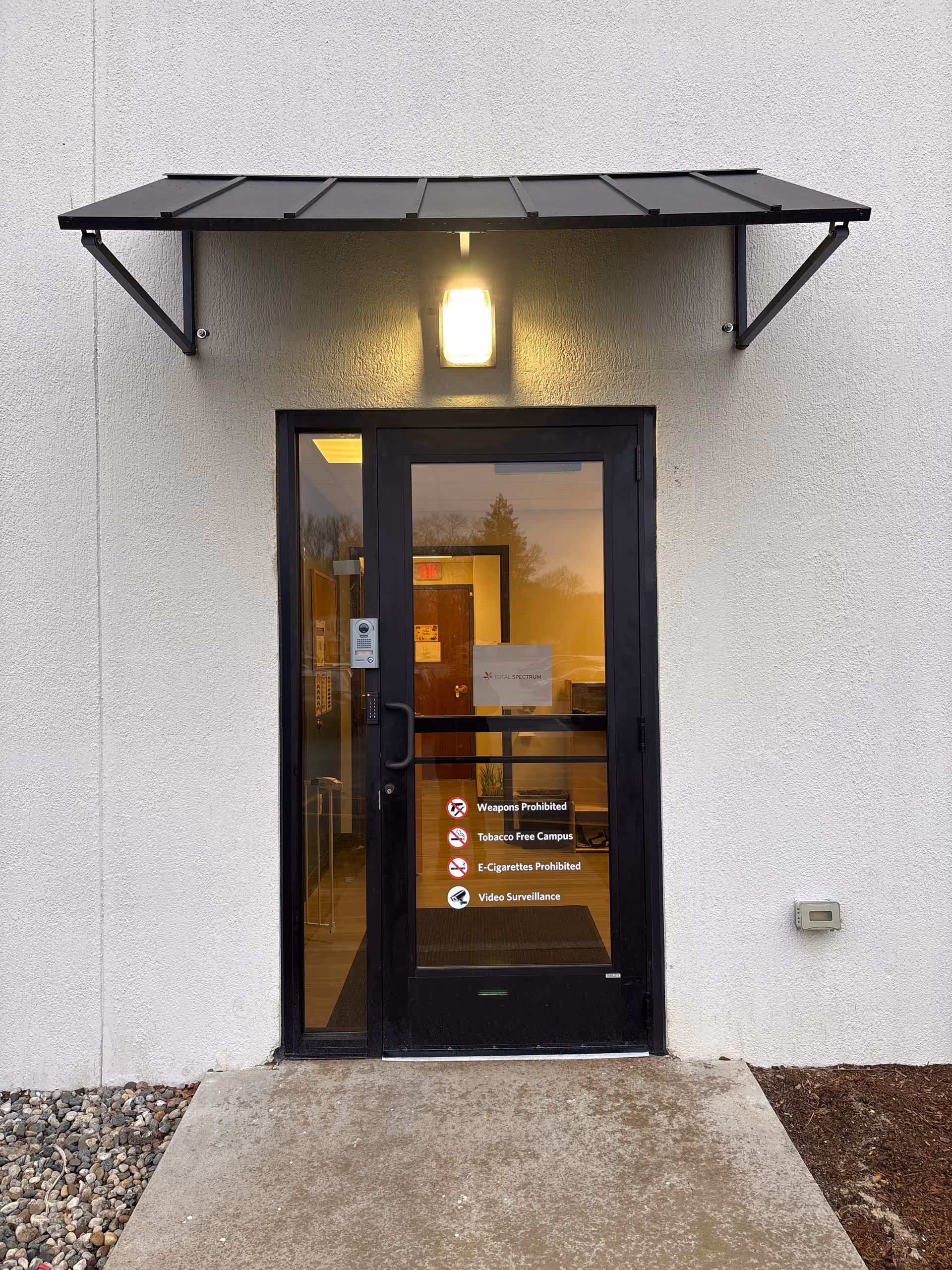 Exterior facility entrance with black door, overhead canopy, and illuminated wall light.