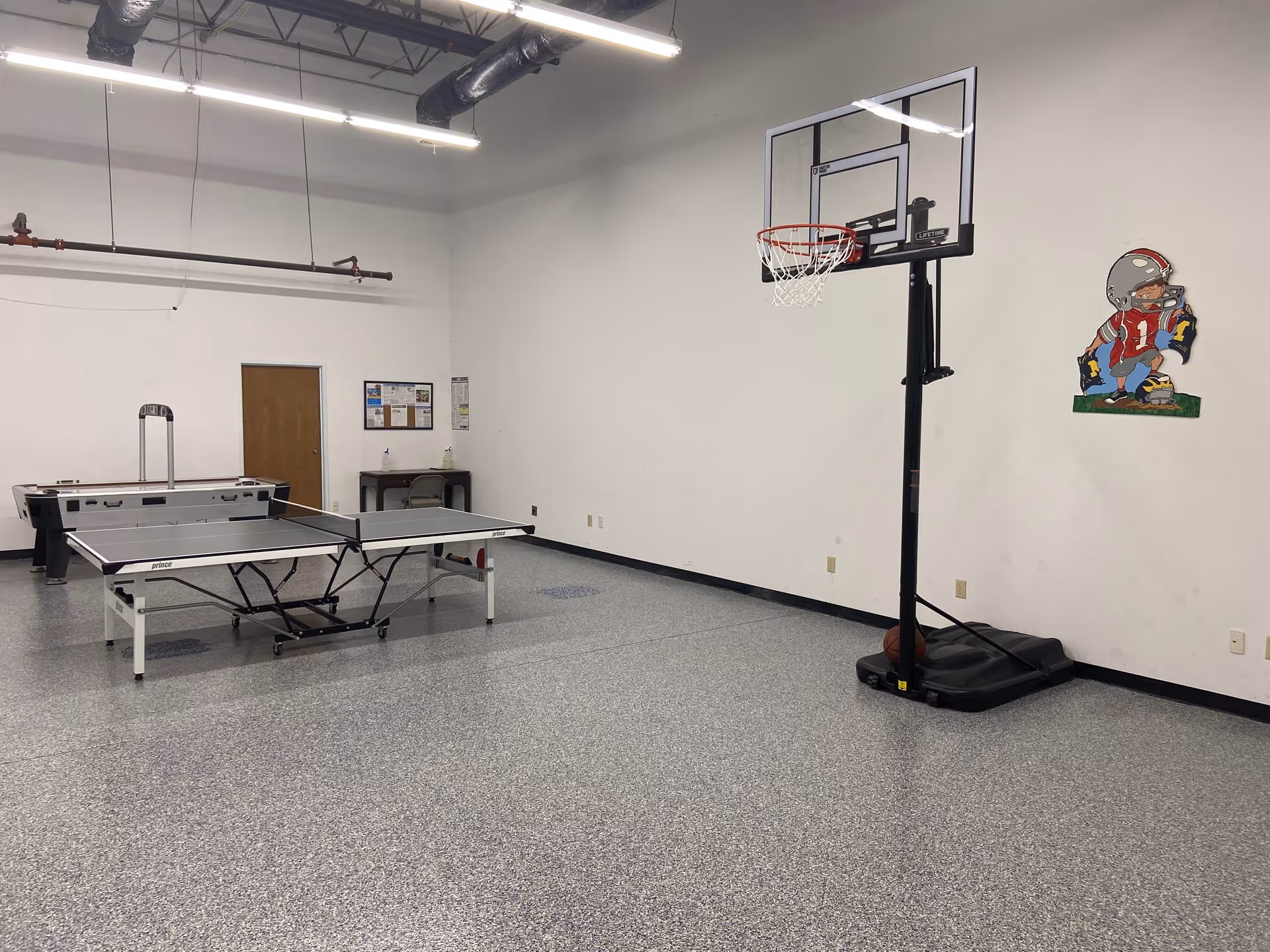 Indoor recreation room with a table tennis setup, basketball hoop, and sports mural.