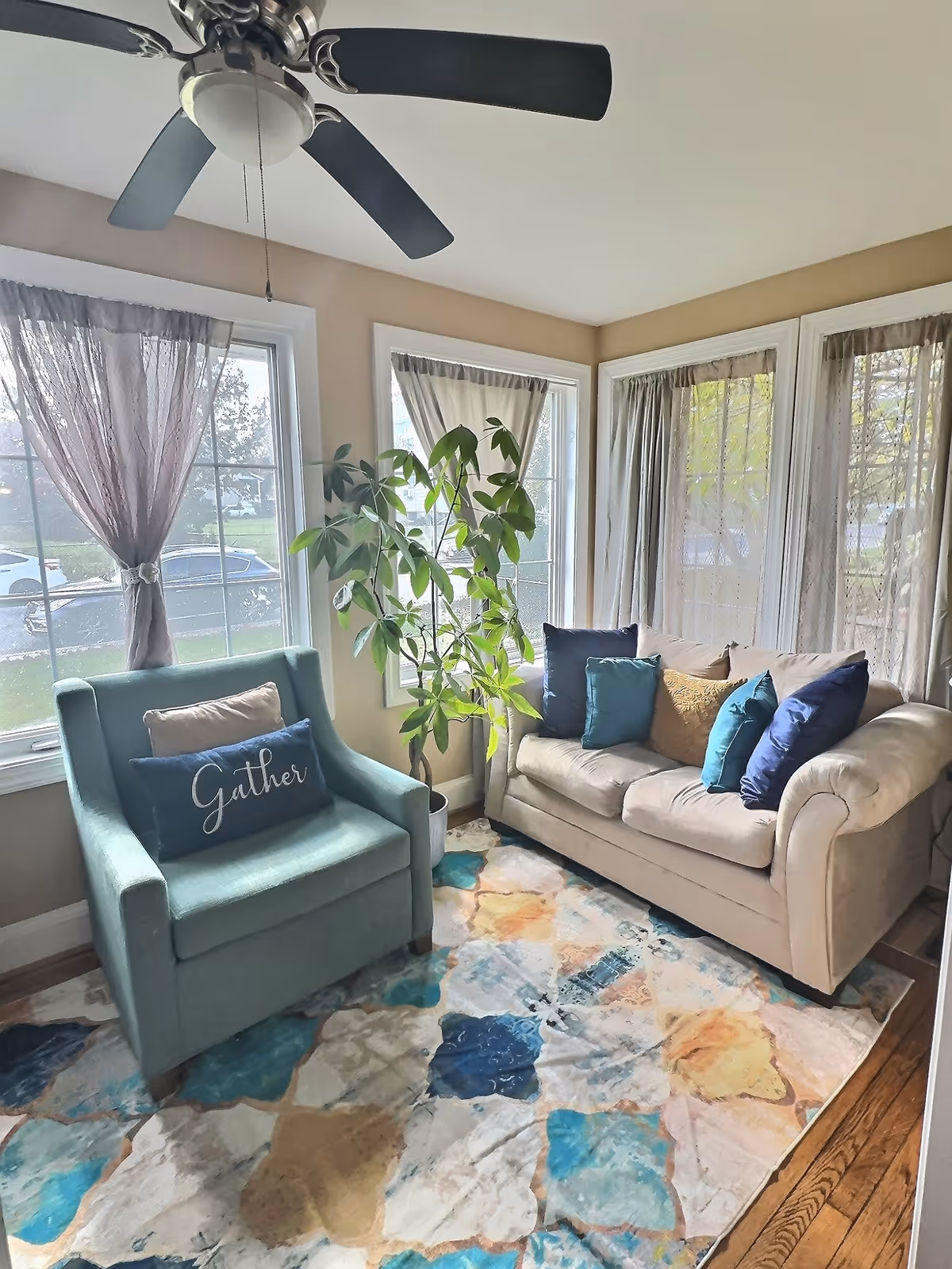 Sunlit living room with teal chair, beige sofa, colorful rug, and potted plant.
