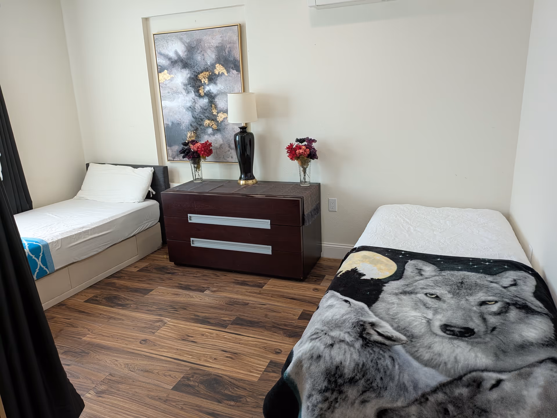 Two beds flank a dark dresser with a lamp and flowers in a tidy bedroom.