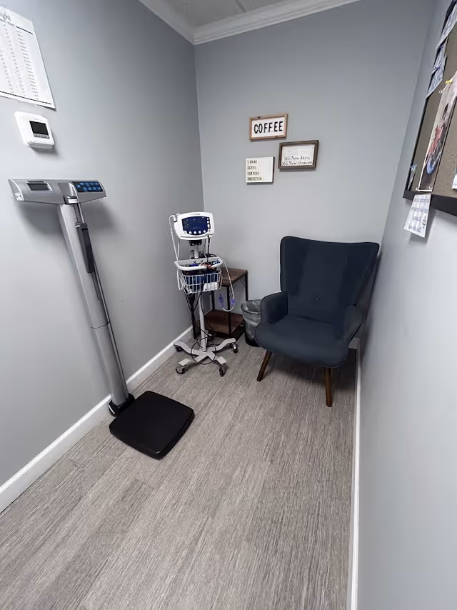 Small medical examination room with a scale, monitor cart, and a blue upholstered chair.